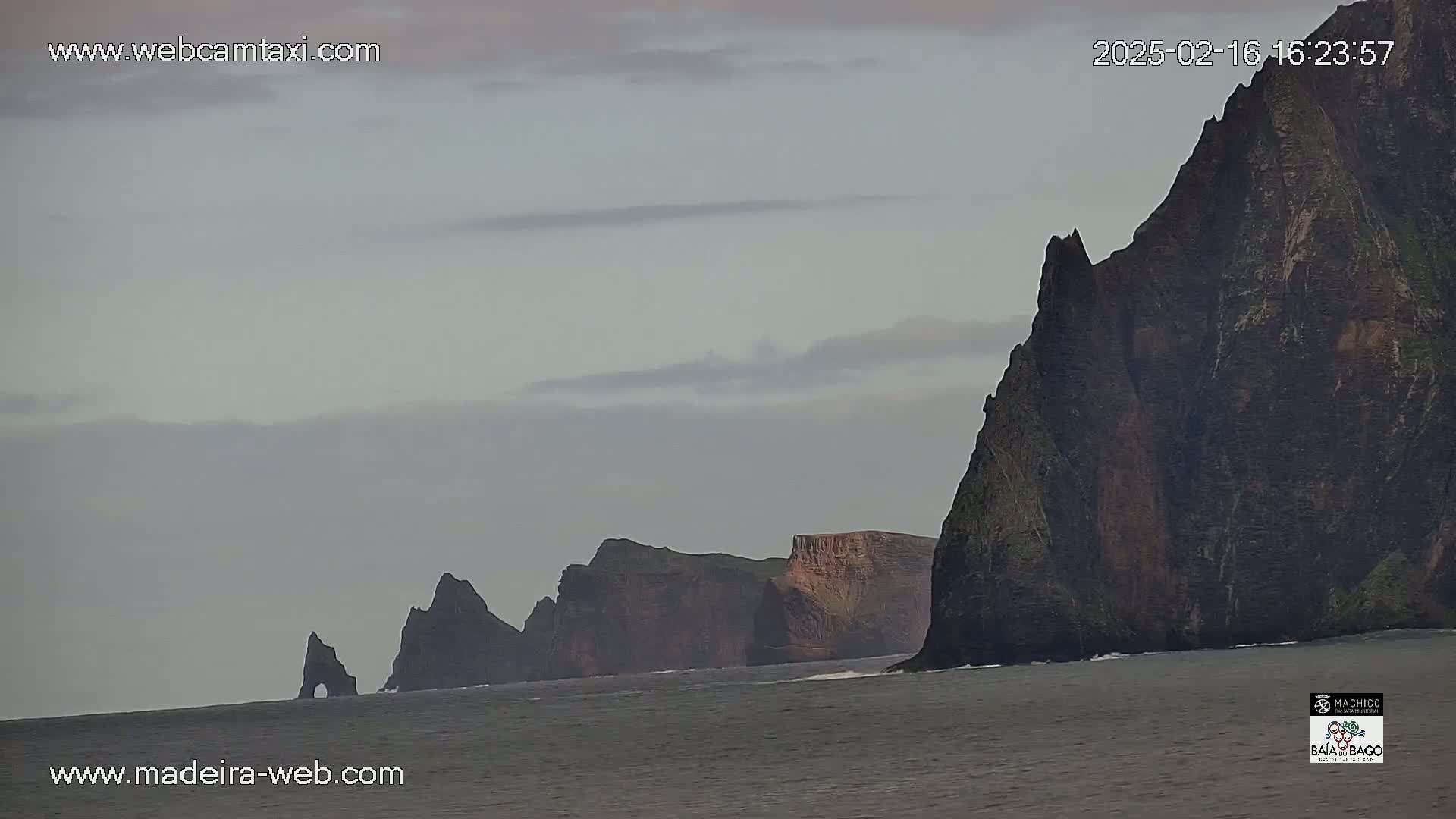 Dark, rocky cliffs rise from a calm, gray ocean under an overcast sky.