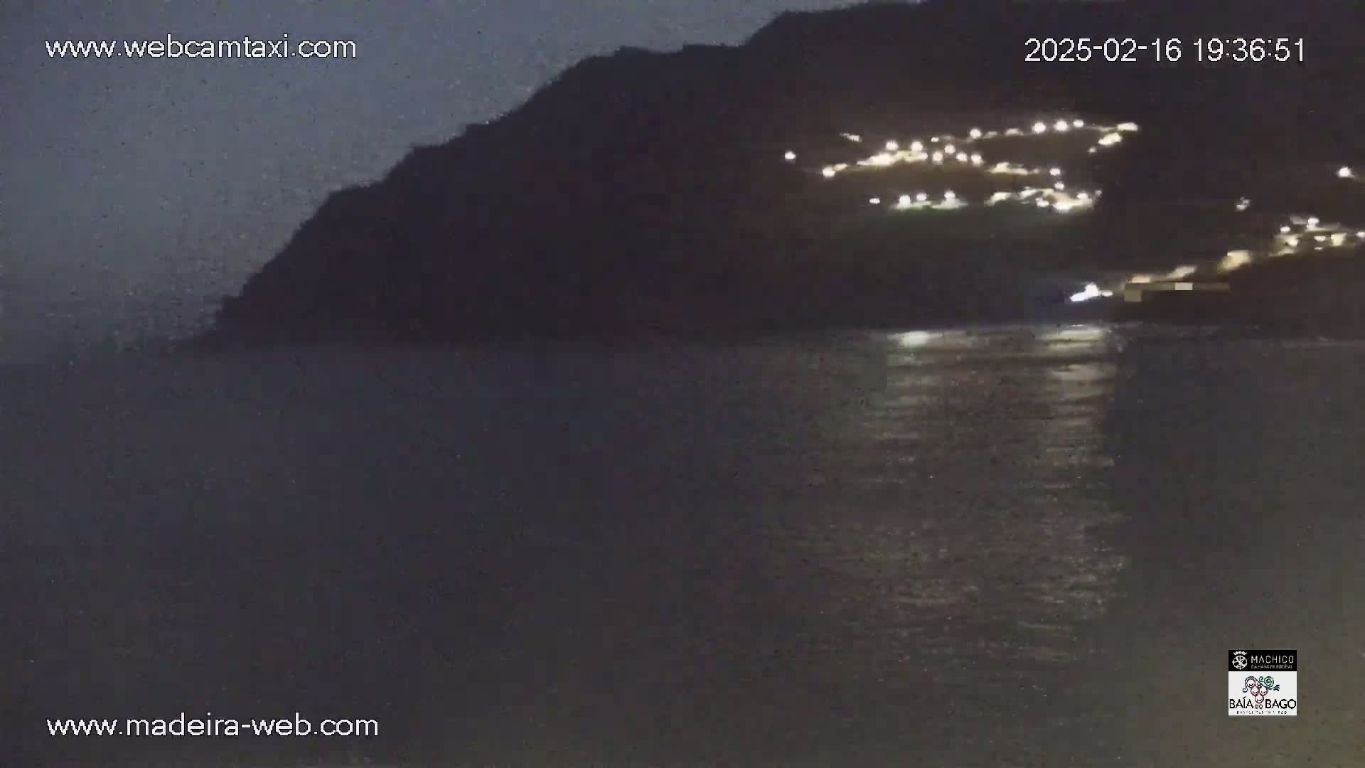 A dark, nighttime scene shows a calm body of water reflecting lights from a hillside settlement on a distant, dark mountain.