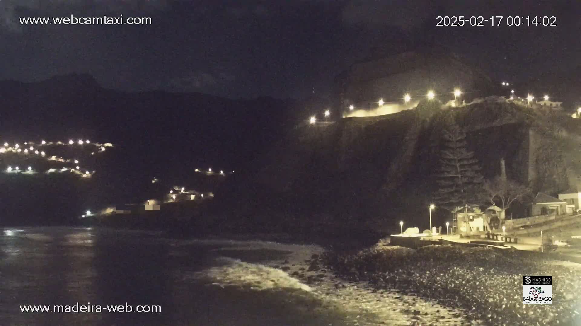 A nighttime coastal scene shows a dark, rocky shoreline with gentle waves, illuminated buildings on a hill, and scattered lights along a cliffside road.