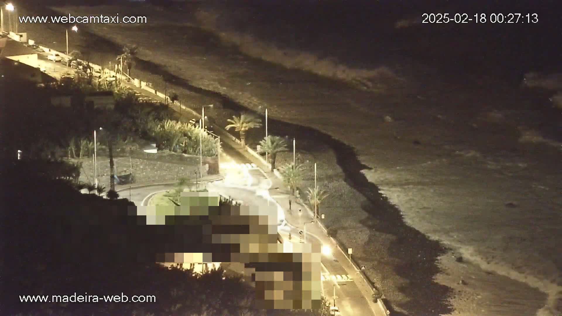 A nighttime, high-angle view shows a dark beach with small waves lapping the shore, next to a roadway with some cars and palm trees, and buildings on a hillside above.