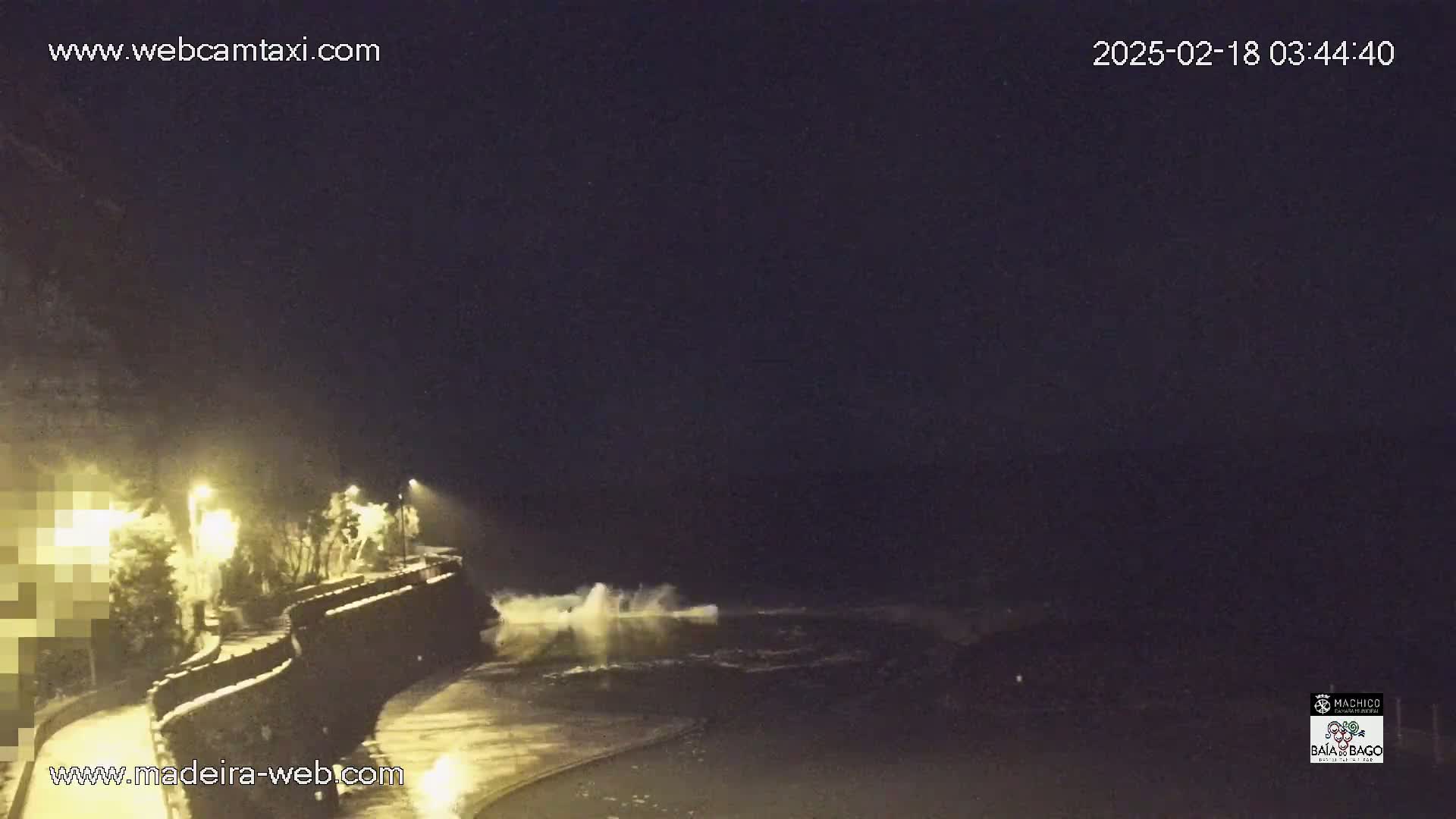 At night, large waves crash against a seawall illuminated by streetlights, under a dark sky.