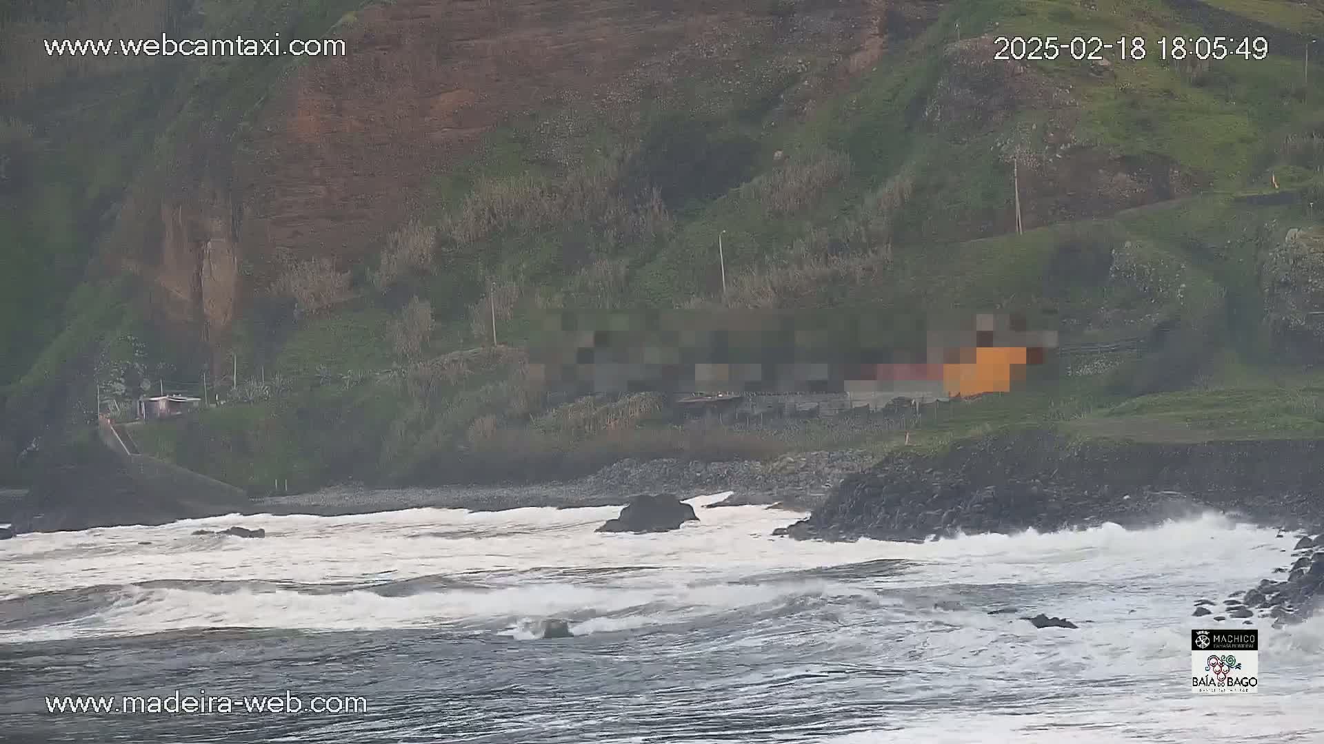 A rocky coast with choppy waves crashes against a dark-colored shore, near a building partially obscured from view, under an overcast sky.