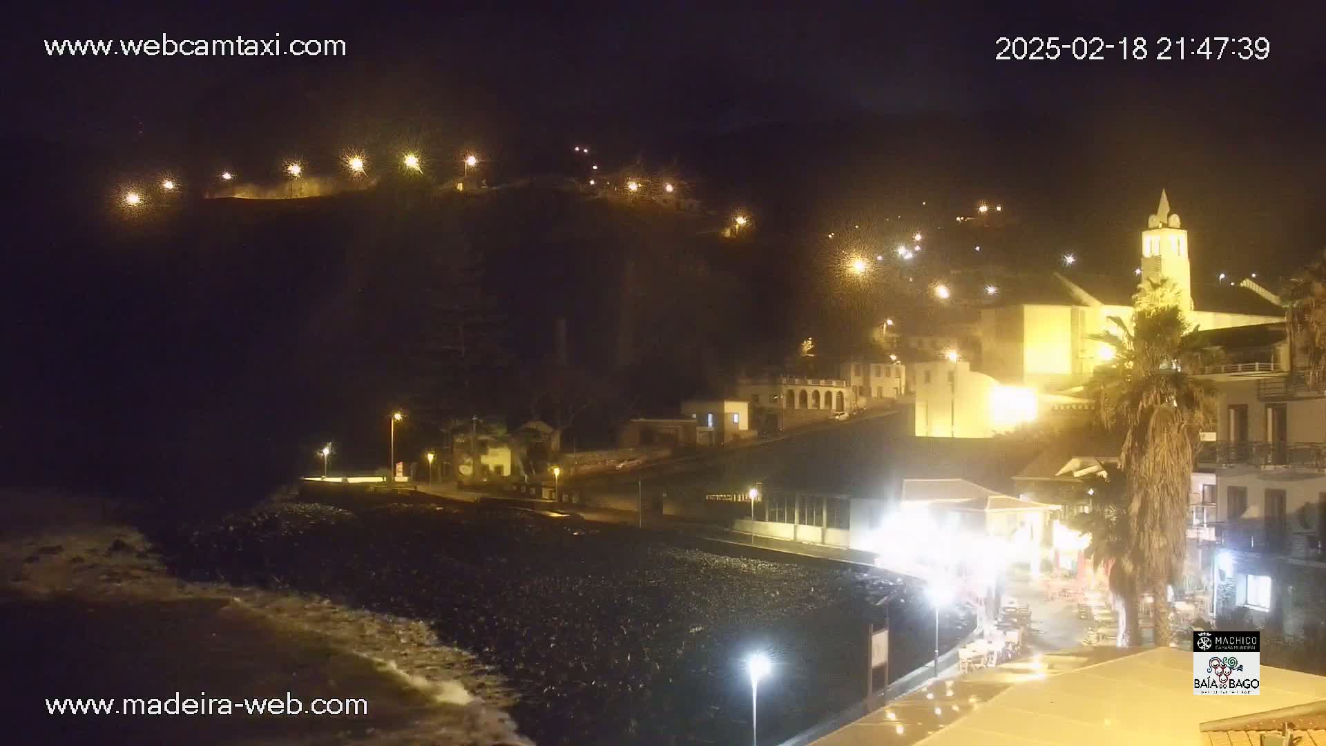 A nighttime coastal town scene with a church, buildings, and palm trees illuminated by streetlights, with some wave action visible on a dark beach.