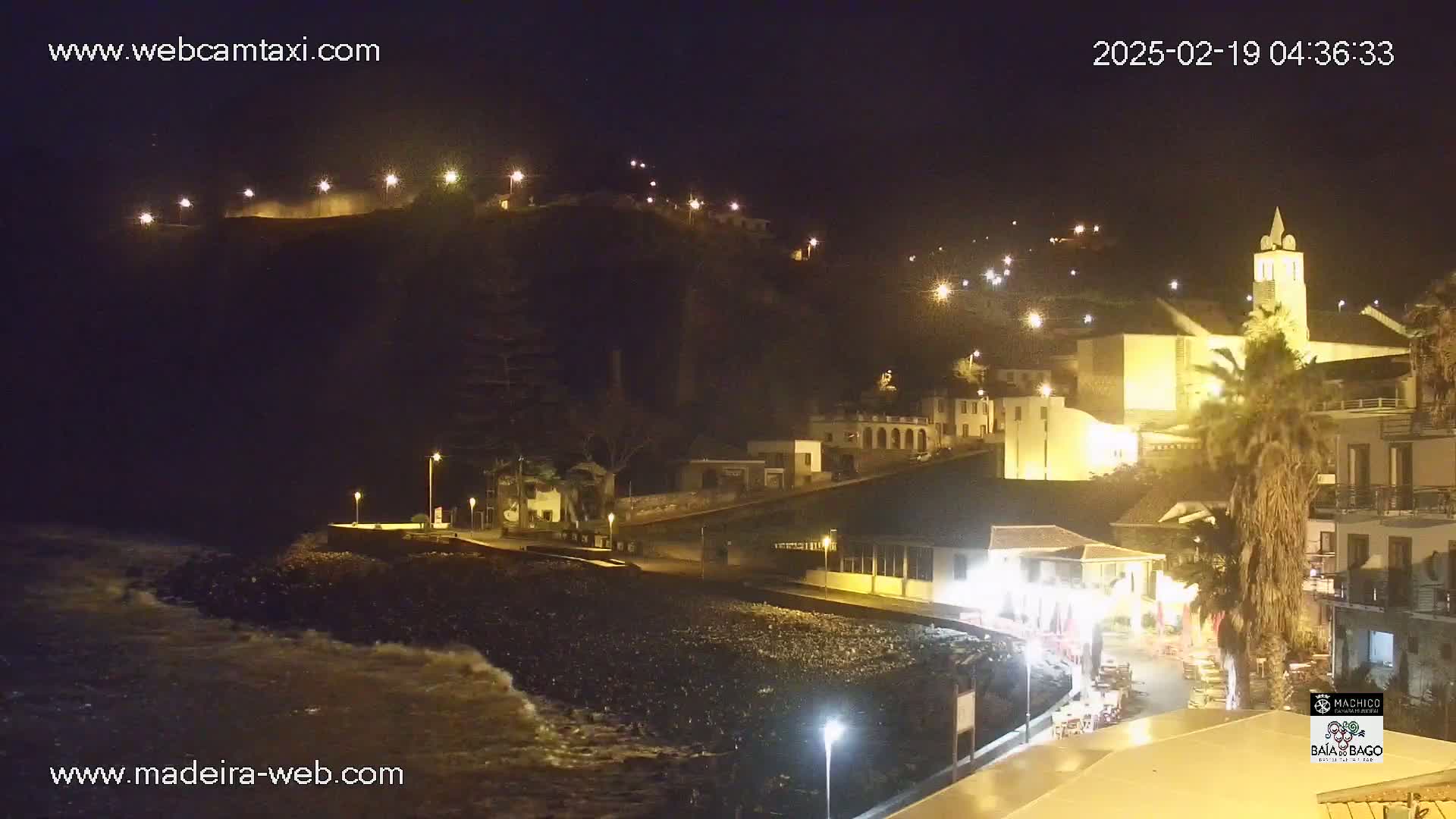 A nighttime coastal town scene shows a rocky beach with gentle waves, illuminated buildings on a hillside, and a church tower.