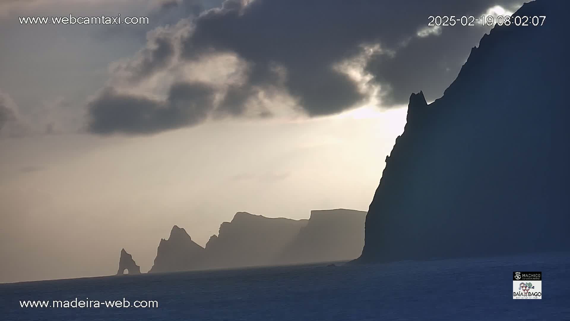 Silhouetted cliffs and rock formations are visible against a cloudy, sunrise sky over calm water.