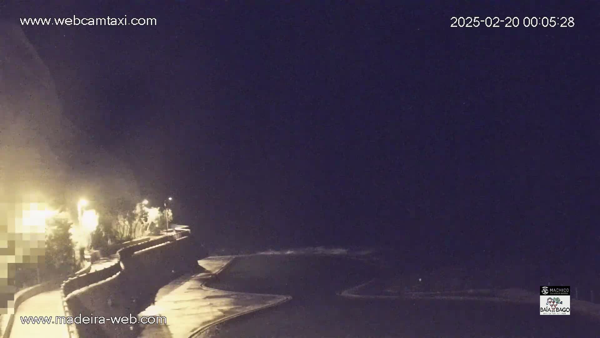 A nighttime coastal scene shows a dark ocean, a curving seawall with lights illuminating a roadway above it, and a dark sky.