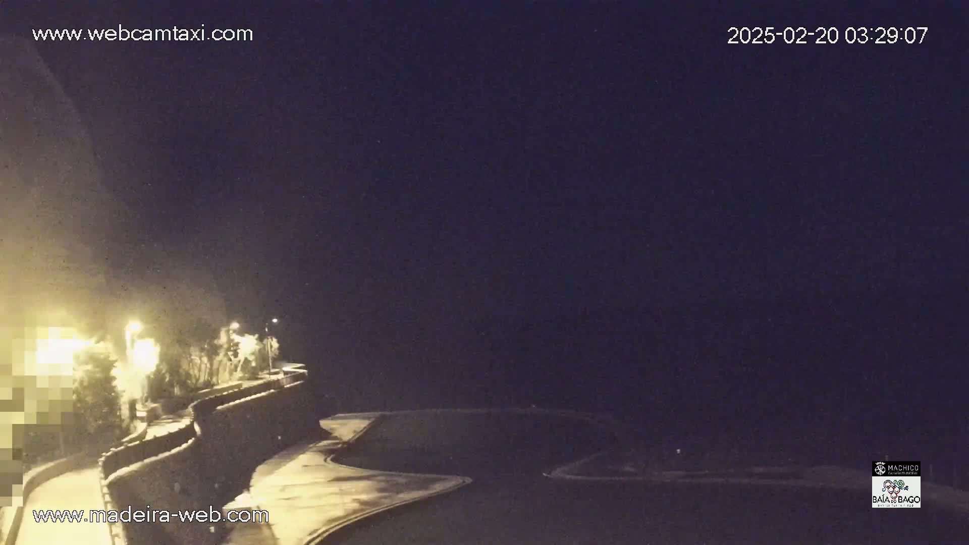 A nighttime, snow-covered road curves along a stone wall, illuminated by streetlights, with a vehicle partially visible in the foreground.