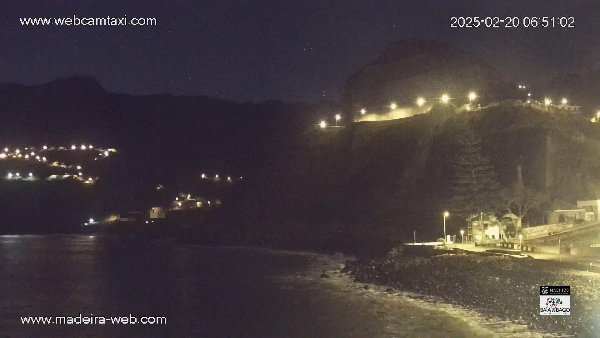 A nighttime coastal scene shows a dark, rocky cliff illuminated by lights, with a small town nestled below, and gentle waves lapping the shore.