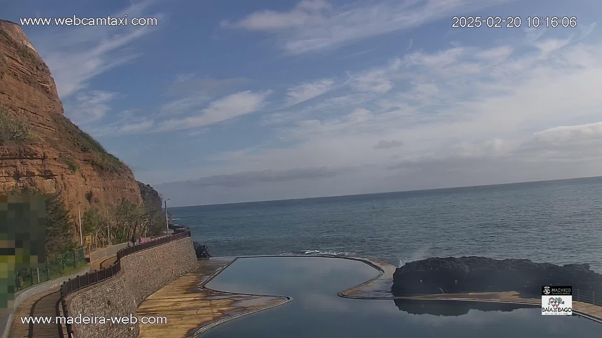A coastal scene on a partly cloudy day shows a rocky cliff, a walkway, and a calm ocean with a man-made pool next to dark volcanic rock formations.