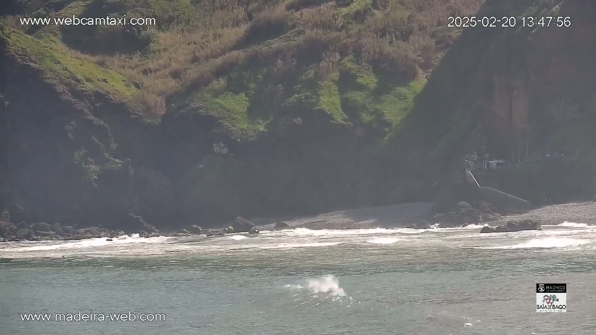 A rocky, grassy cliff overlooks a pebbly beach with gentle waves on a sunny day.