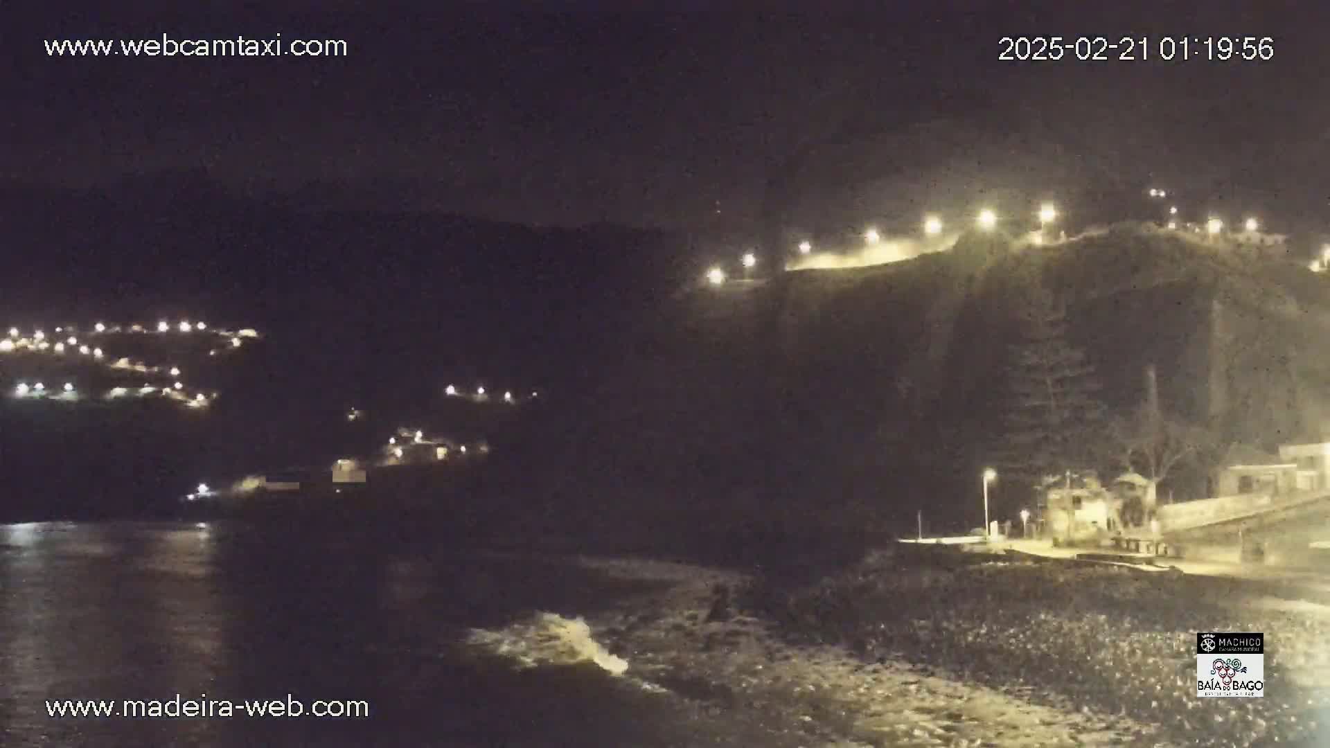 A nighttime coastal scene shows a dark ocean with waves lapping a rocky shore,  illuminated buildings on a hillside, and lights along a clifftop road.