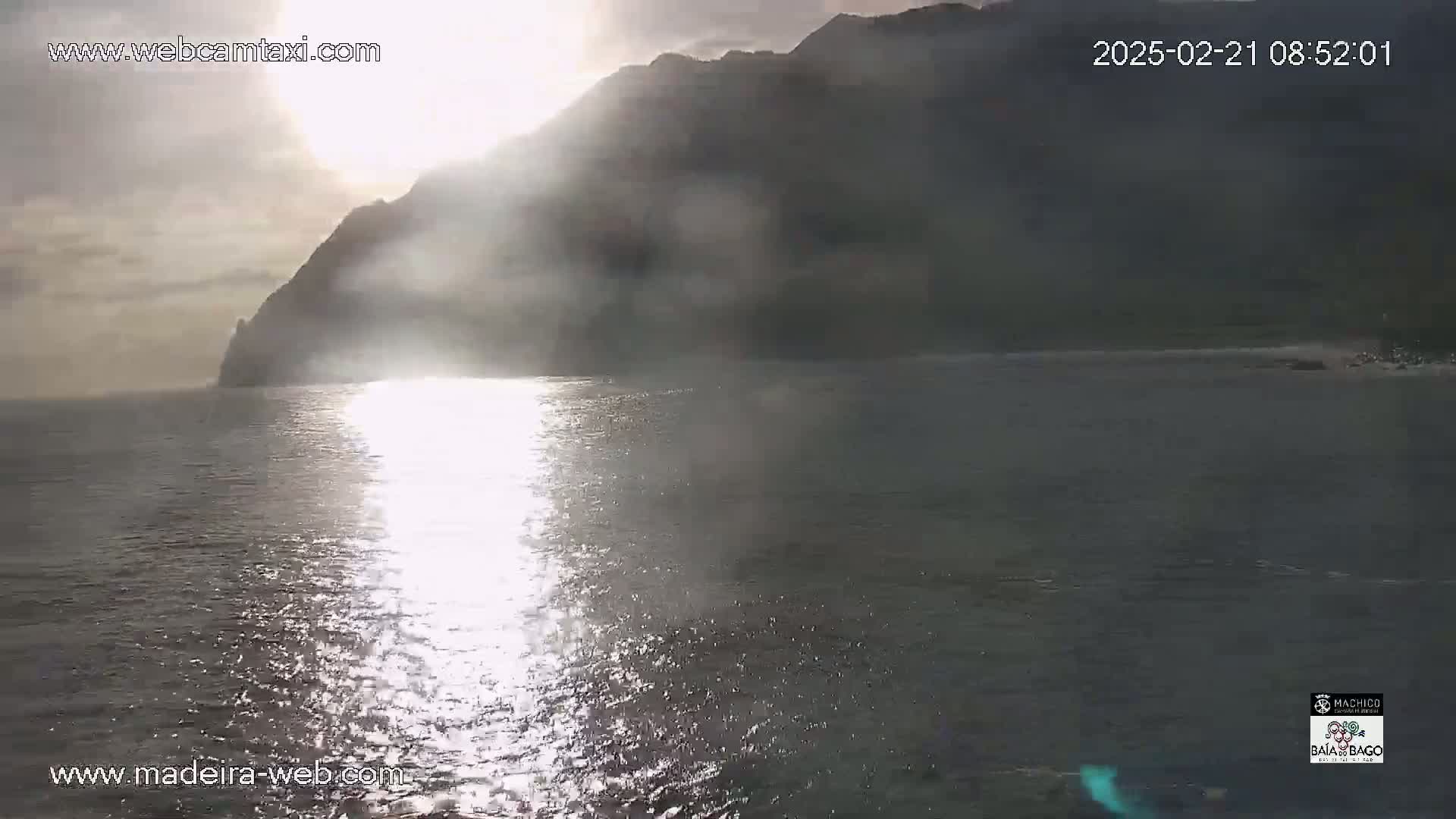 A sunlit, hazy scene shows a calm body of water reflecting the sunlight, with a dark mountain silhouetted against a cloudy sky in the background.
