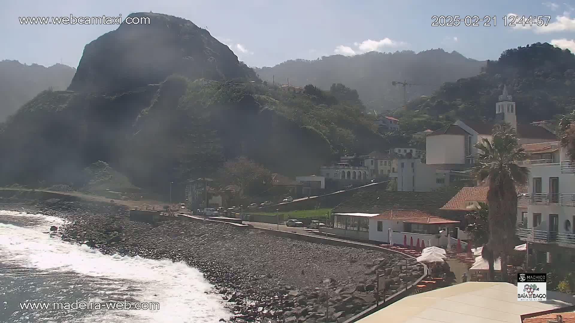 A rocky beach with waves crashing against the shore, backed by a village nestled against a hazy mountainside under a mostly clear sky.