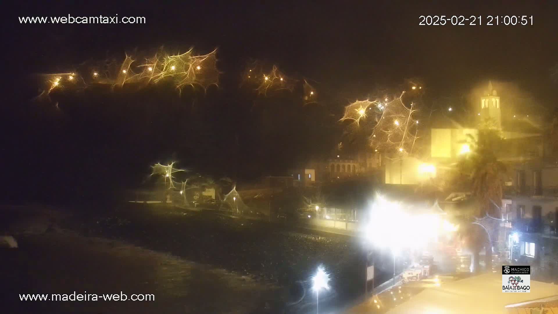 A nighttime coastal town scene with illuminated buildings and streets, under foggy or rainy conditions, shows what appear to be light streaks in the sky.