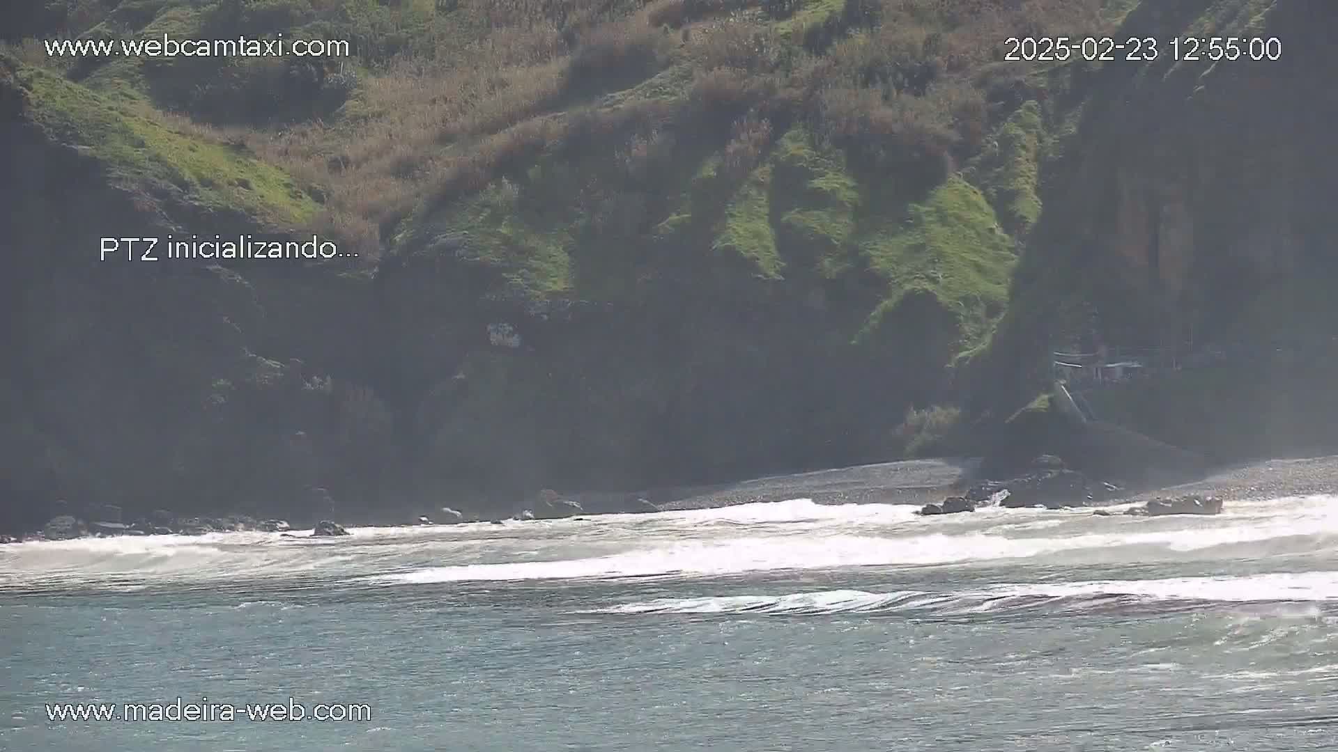 Ocean waves break on a rocky shore beneath a green cliff on a sunny day.