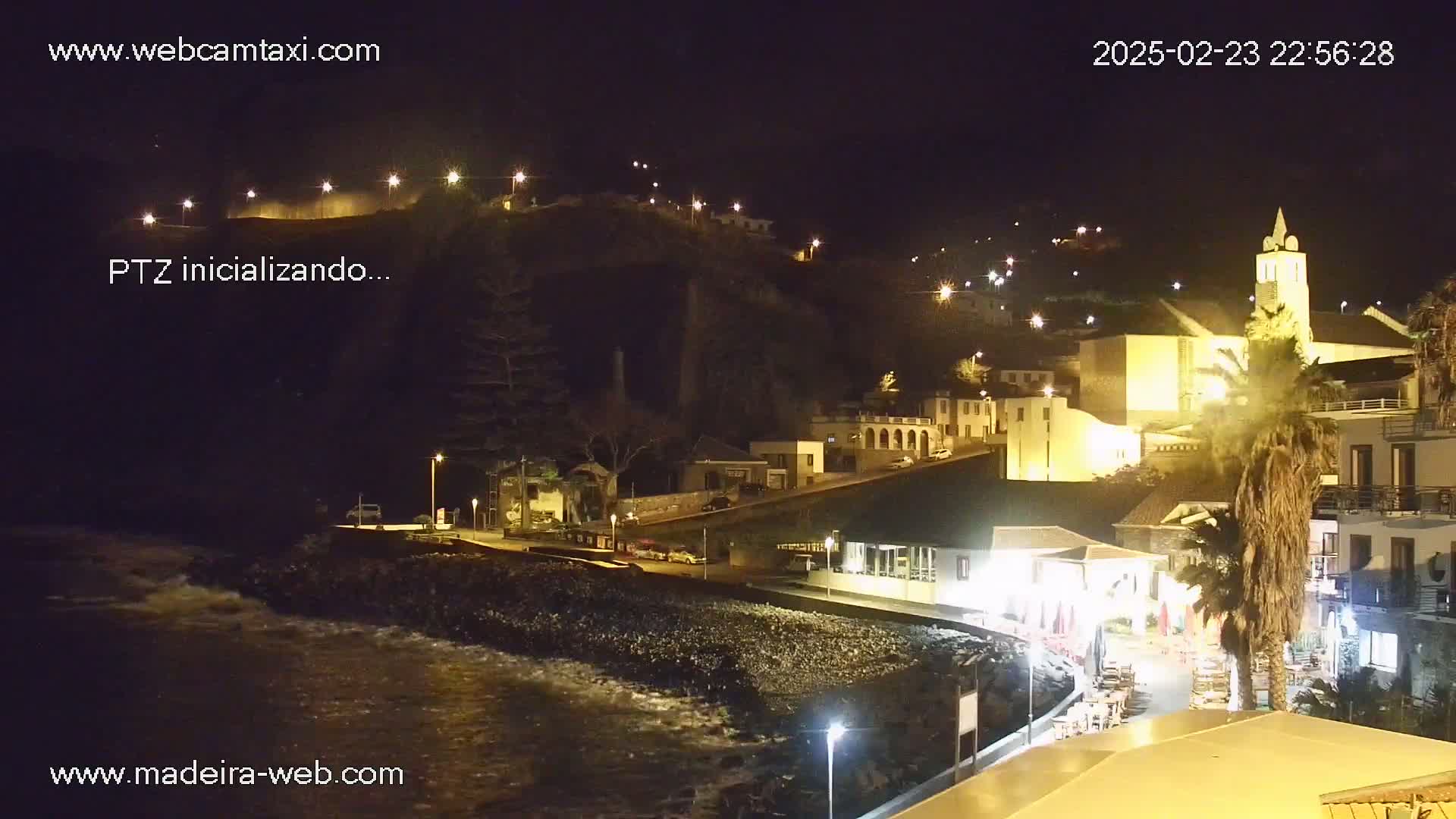 A nighttime coastal town scene with a church and buildings illuminated, gentle waves lapping at a rocky shore.