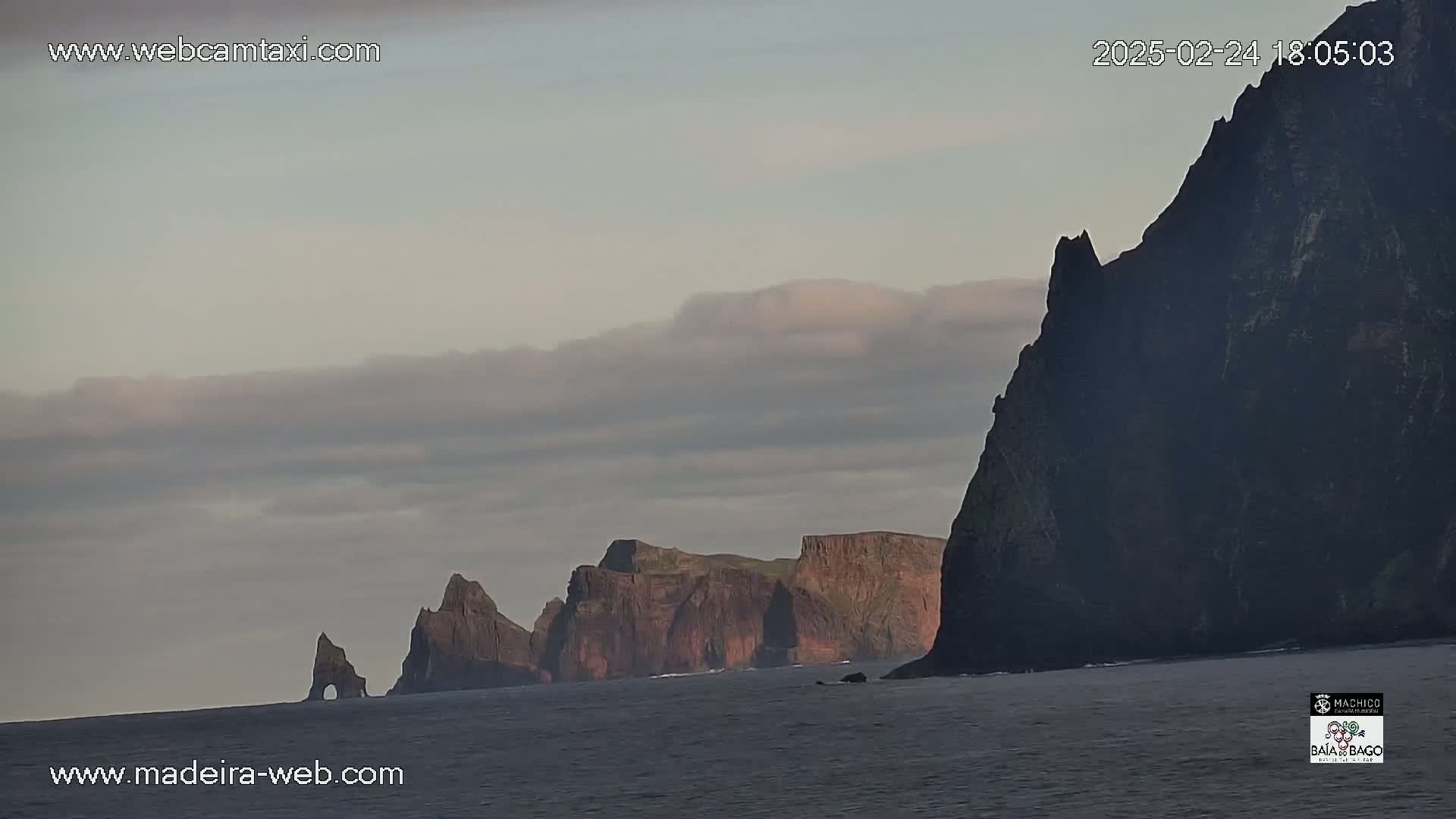 Dark, rocky cliffs rise from a calm ocean under a cloudy, overcast sky.