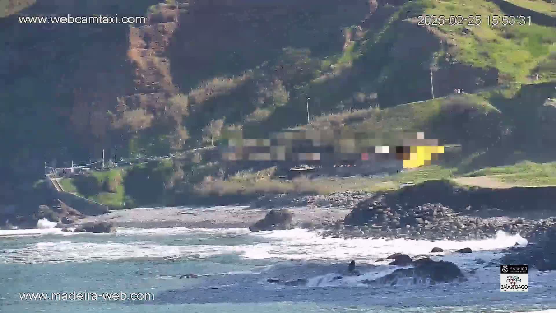 A rocky coastline with small waves crashing against dark rocks and a building on a hillside overlooking the ocean.
