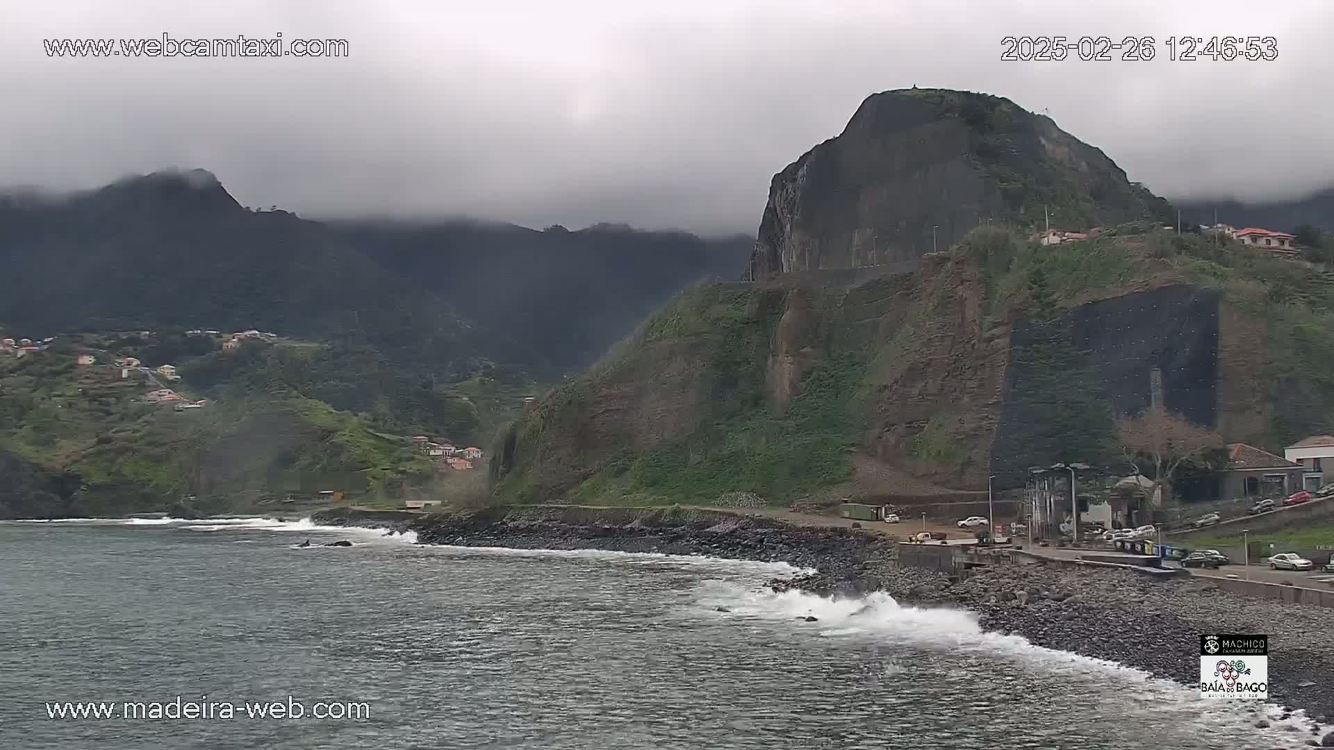 A rocky coastline with a village and a large cliff face is shown under an overcast sky, with waves crashing on the shore.