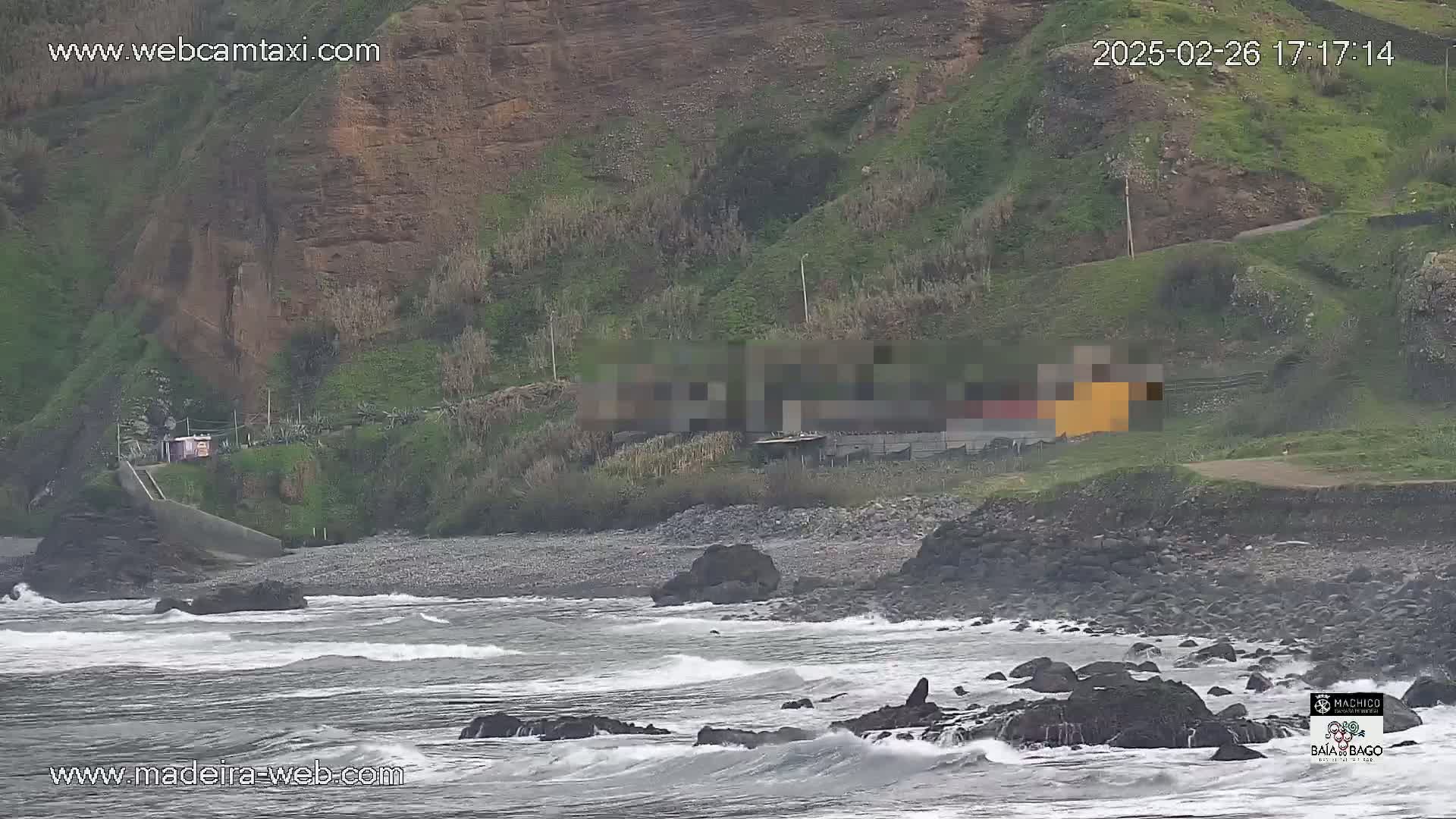 A rocky coastline with small waves crashes against dark rocks and a pebble beach, next to green hillsides under an overcast sky.