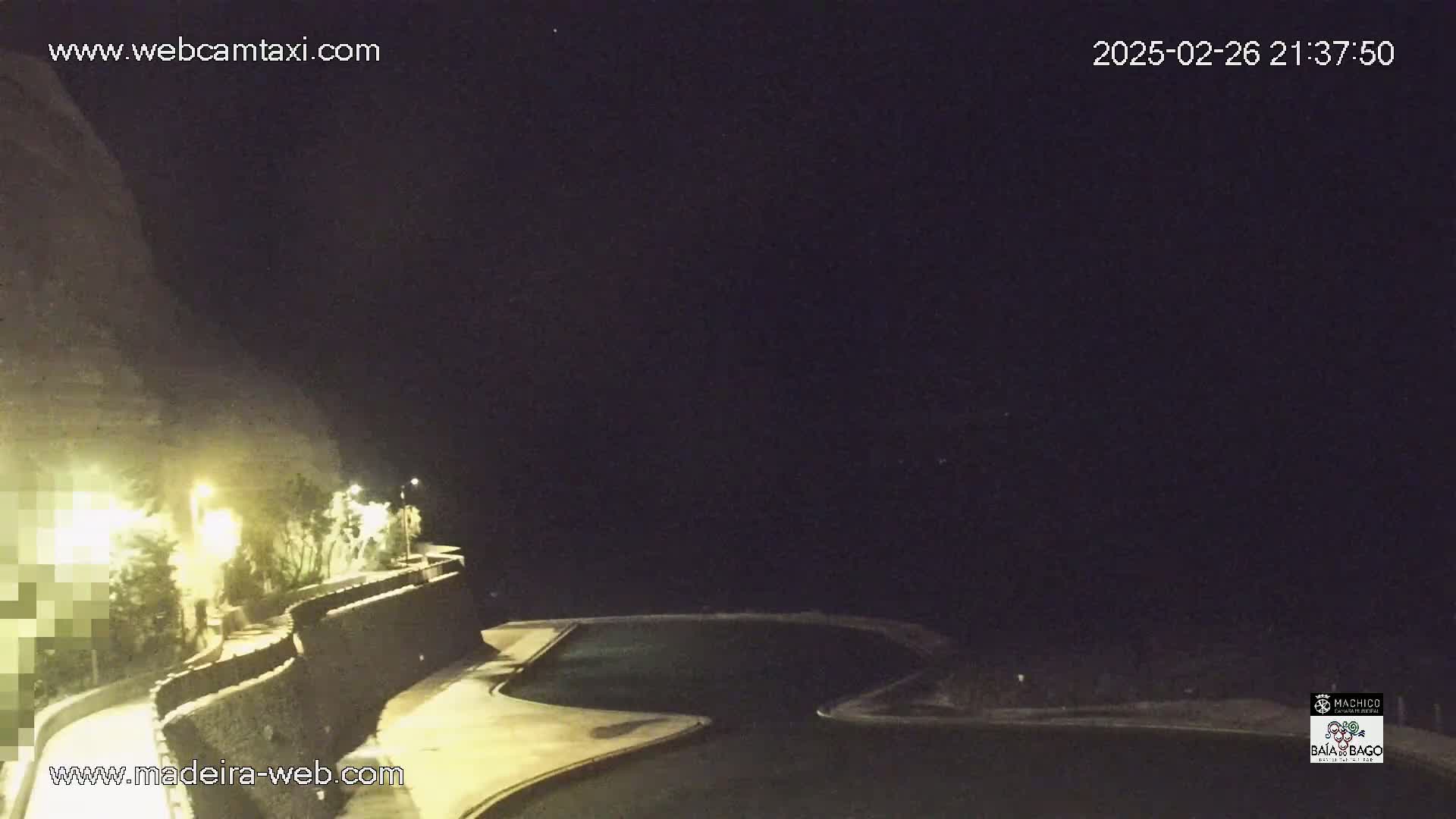 A nighttime, outdoor view shows a dark, cliff-like hill overlooking a pool area lit by artificial lights.