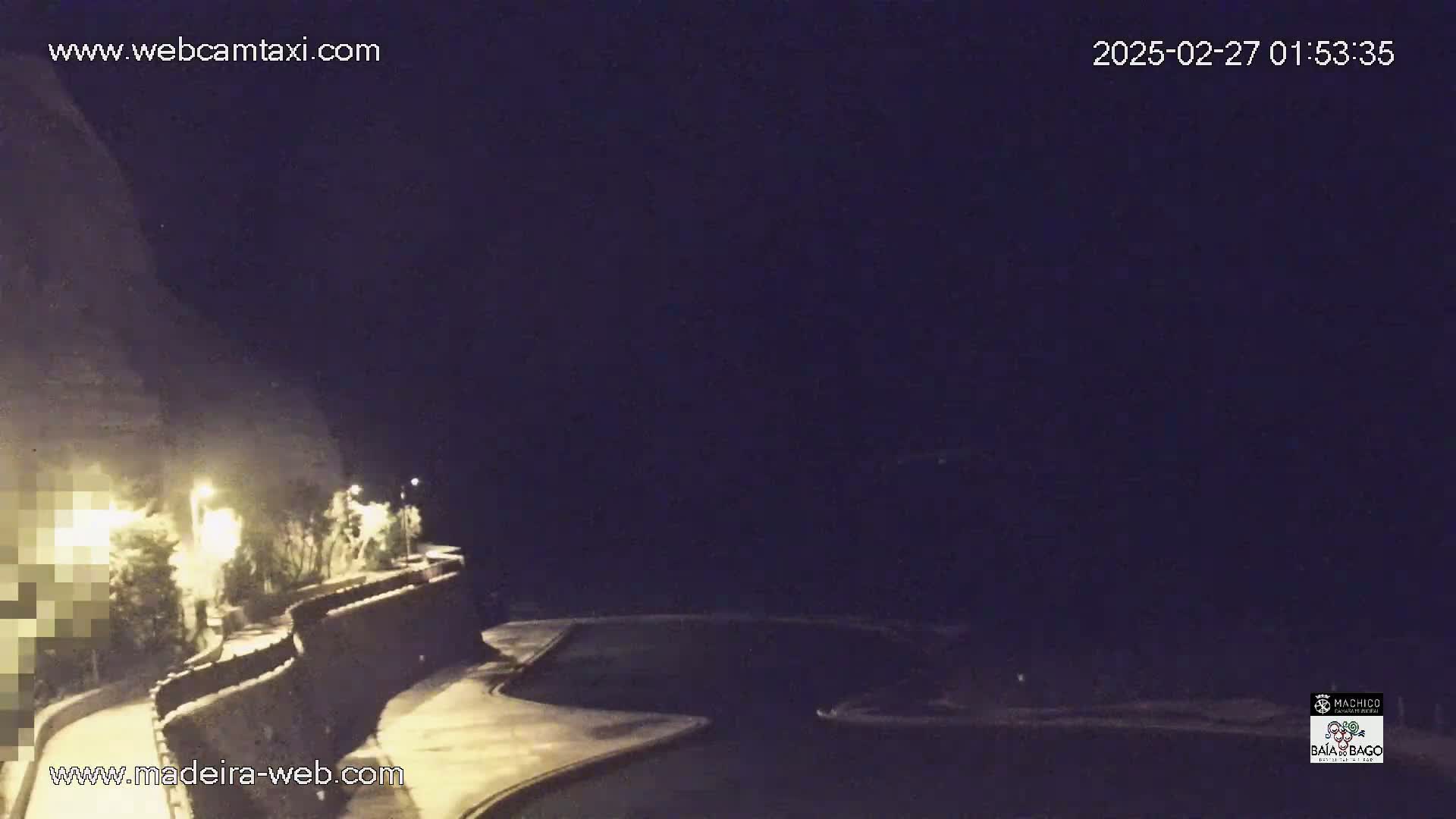 A nighttime, dimly lit view of a roadway curving alongside a low stone wall, with vegetation and streetlights visible.