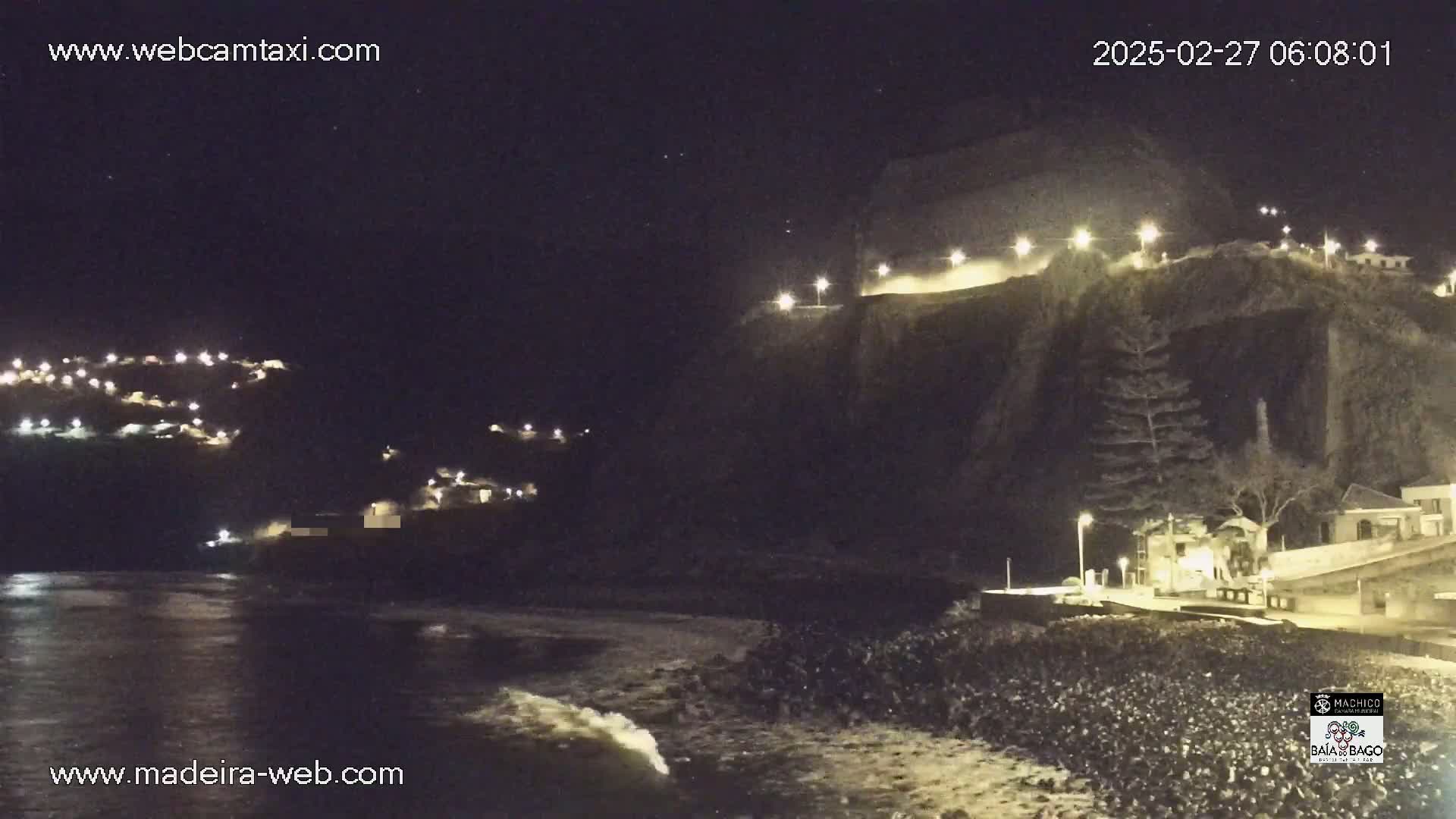 A nighttime coastal scene shows dark cliffs with illuminated buildings and a rocky shore with gentle waves under a clear, starry sky.