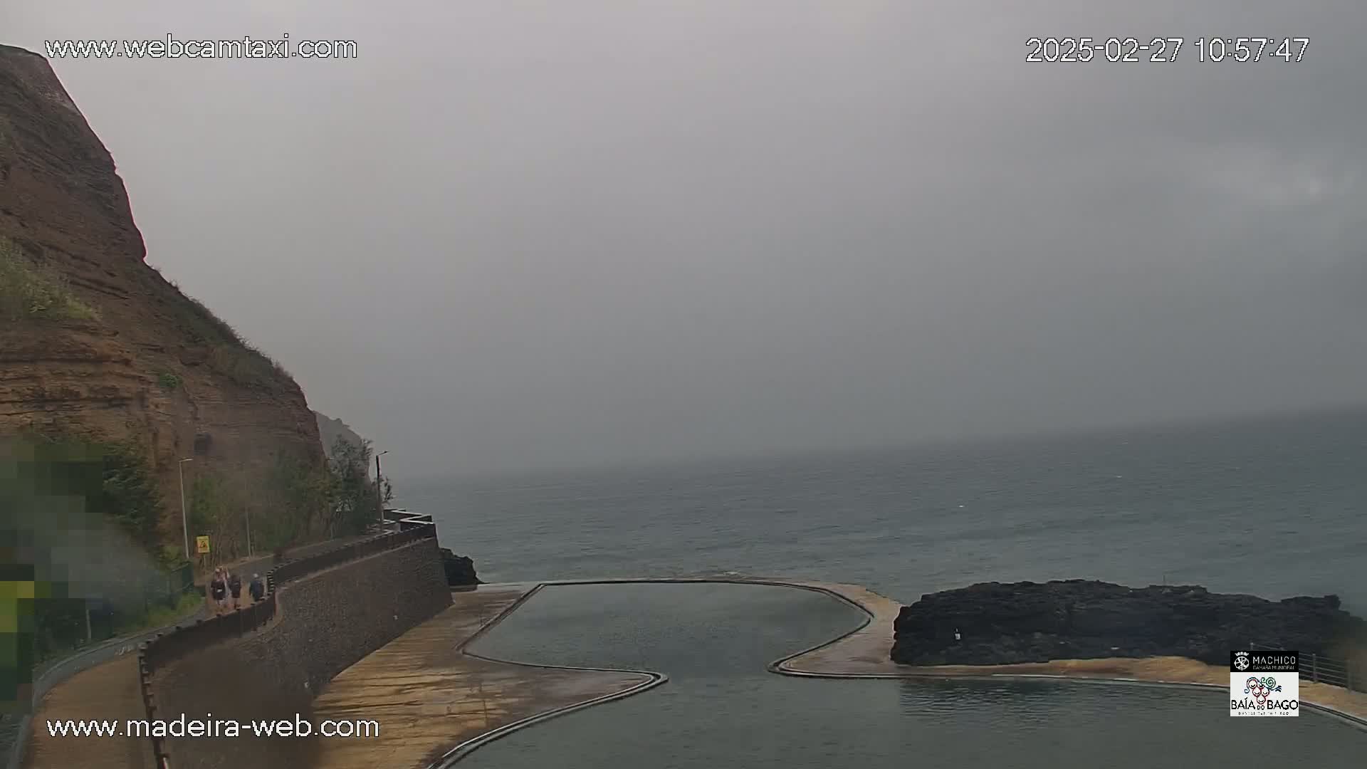 A rocky cliff overlooks a calm ocean and a serpentine-shaped pool on an overcast day.