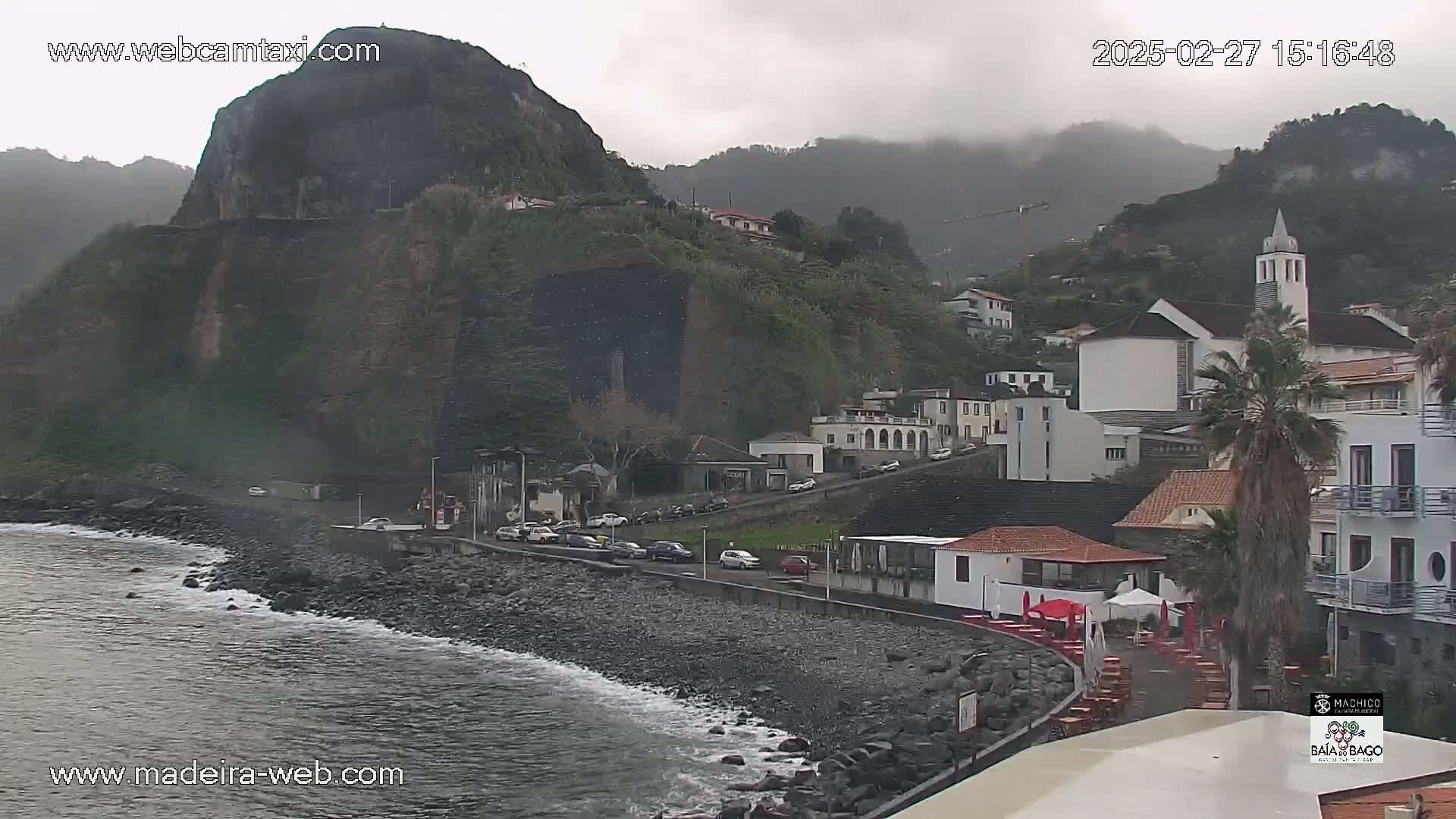 A coastal town nestled against a steep, rocky hillside under an overcast sky, with a dark-colored ocean lapping at a rocky shore.