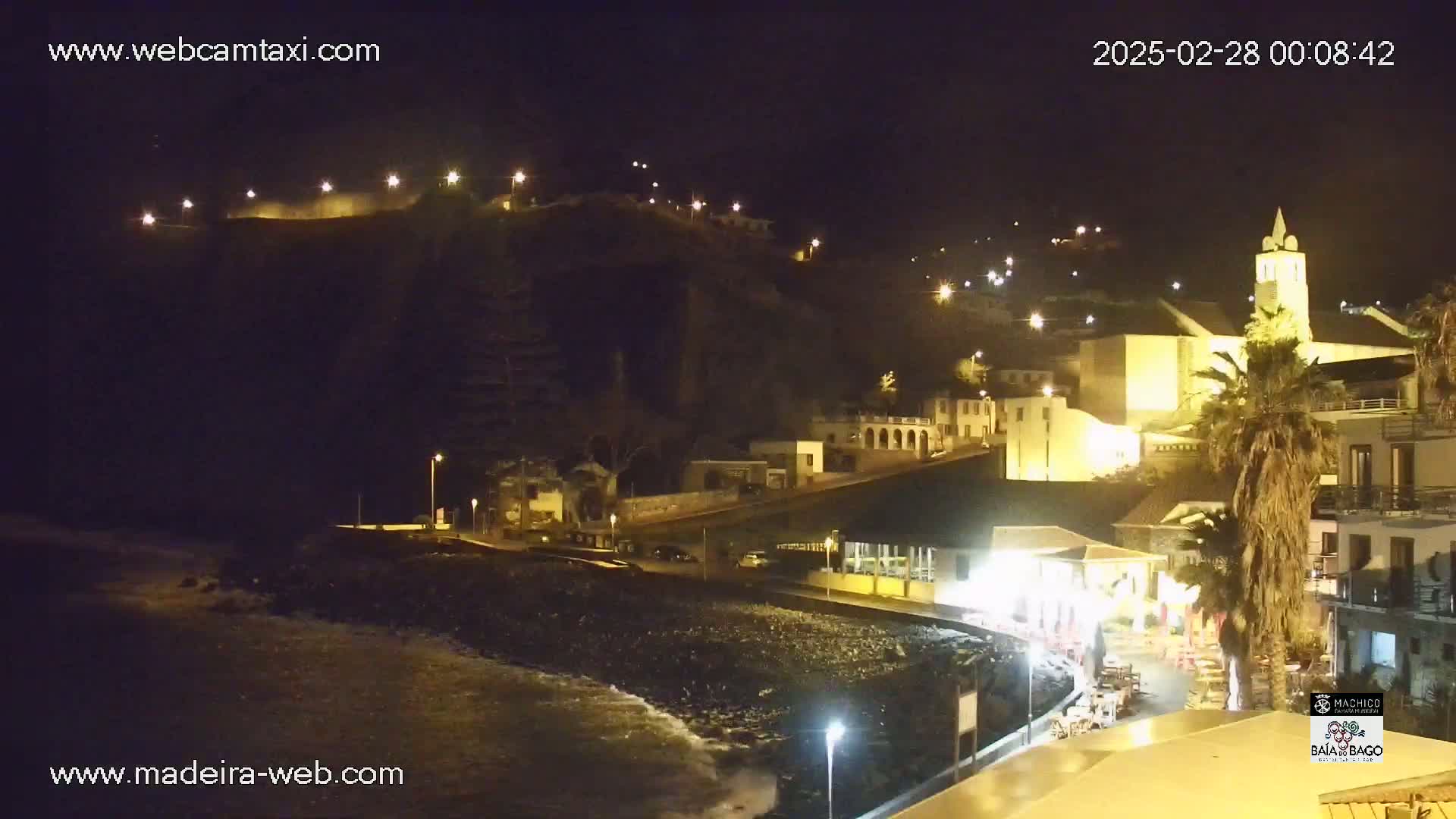 A nighttime coastal town with buildings lit up on a hillside overlooking a dark, rocky beach with gentle waves.