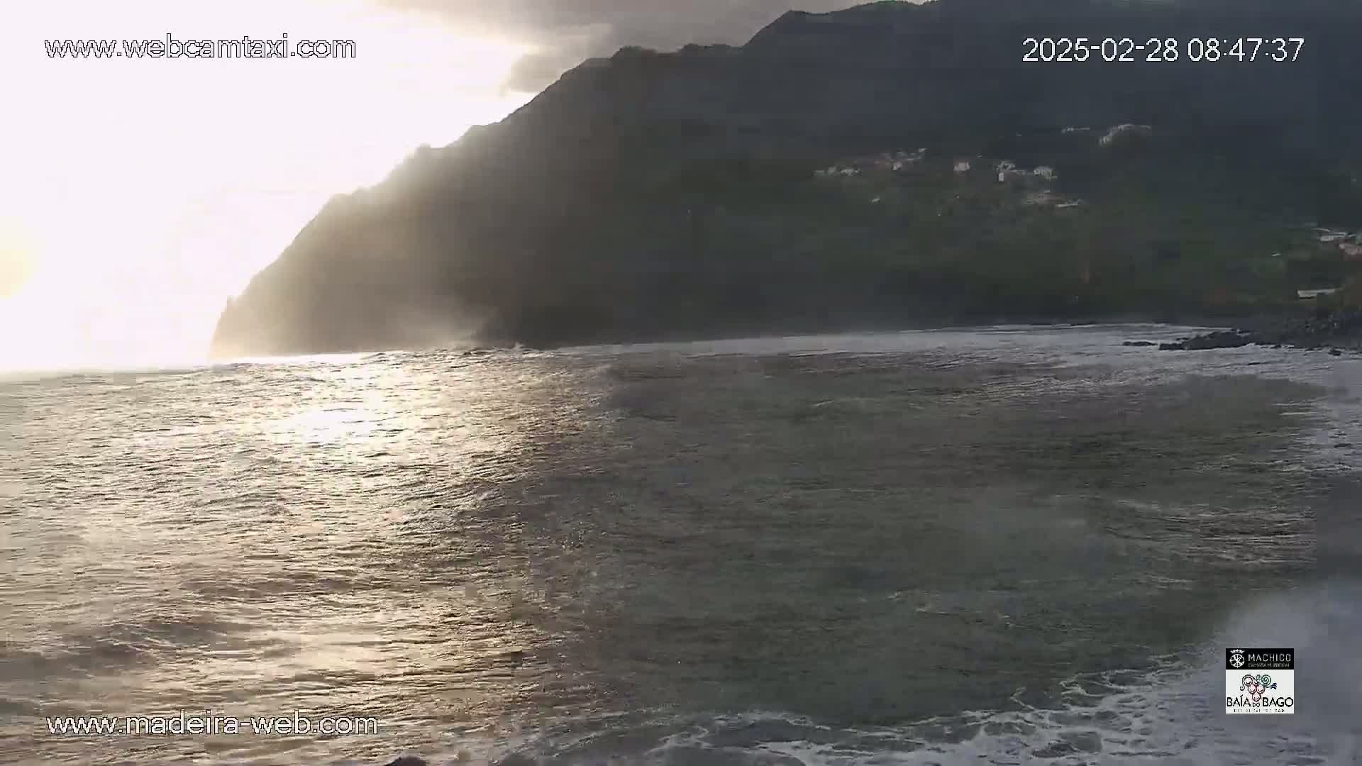 A dark, rocky cliff face is partially illuminated by the sun, overlooking a choppy ocean with small waves.