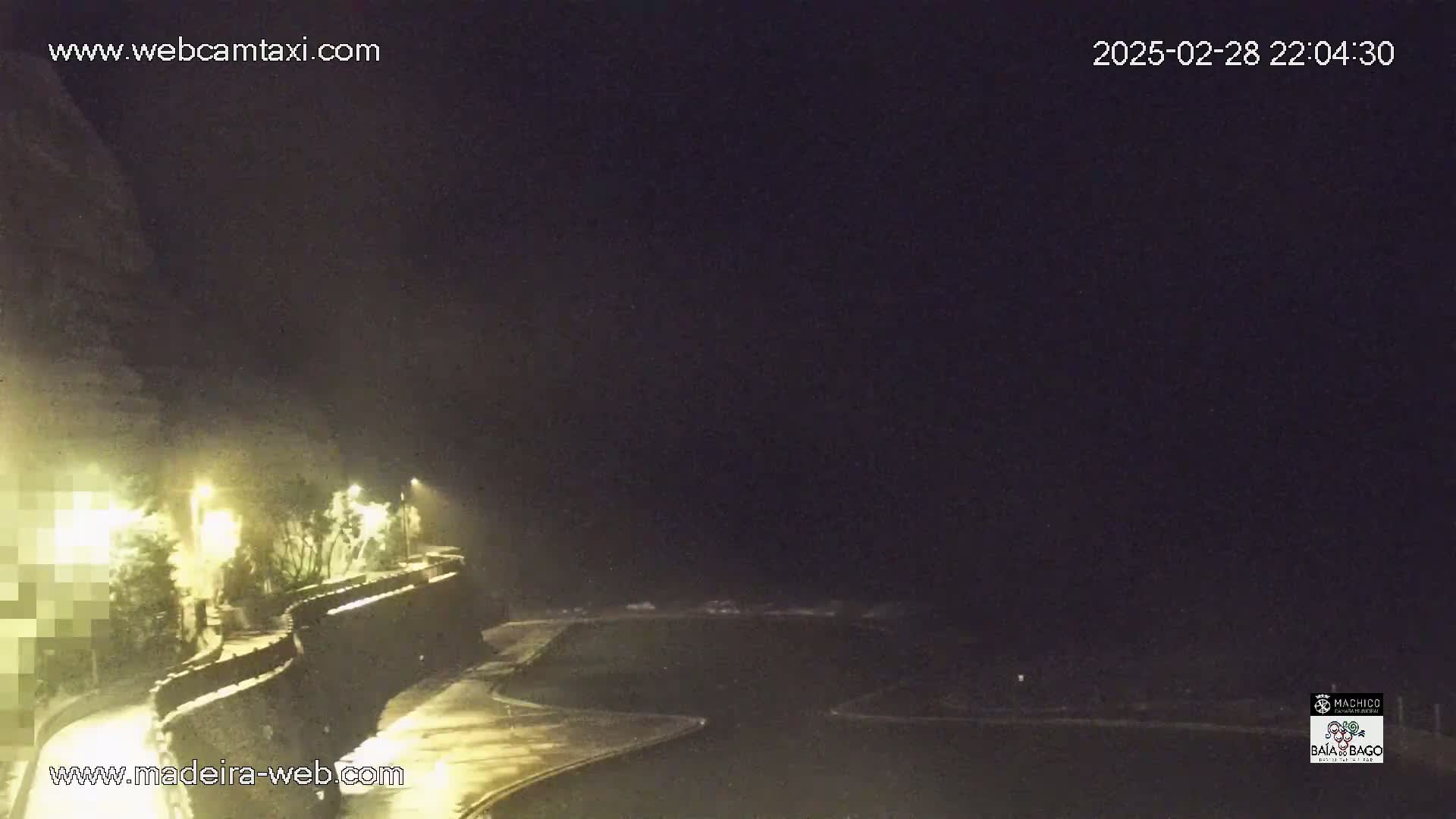 A nighttime view of a coastal road illuminated by streetlights, with a dark sky and possibly some fog or mist.