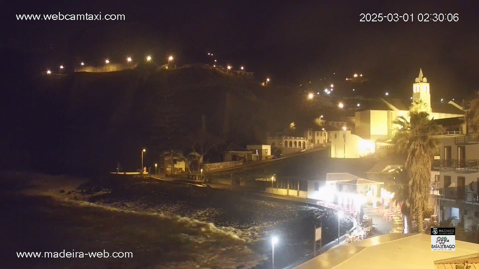 A nighttime coastal town scene shows a dark beach with waves lapping the shore, illuminated buildings on a hillside, and a prominent church tower.