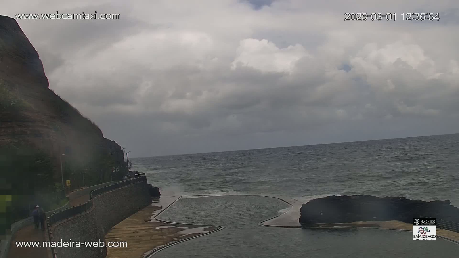 A cloudy day shows a rocky cliff overlooking an ocean with waves crashing near a man-made pool.