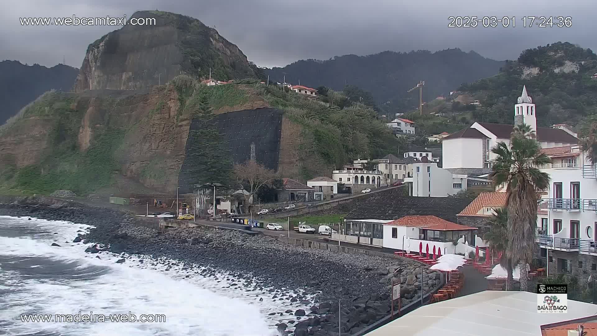 A coastal town nestled against a steep cliffside under an overcast sky, with waves crashing on a dark, rocky shore.