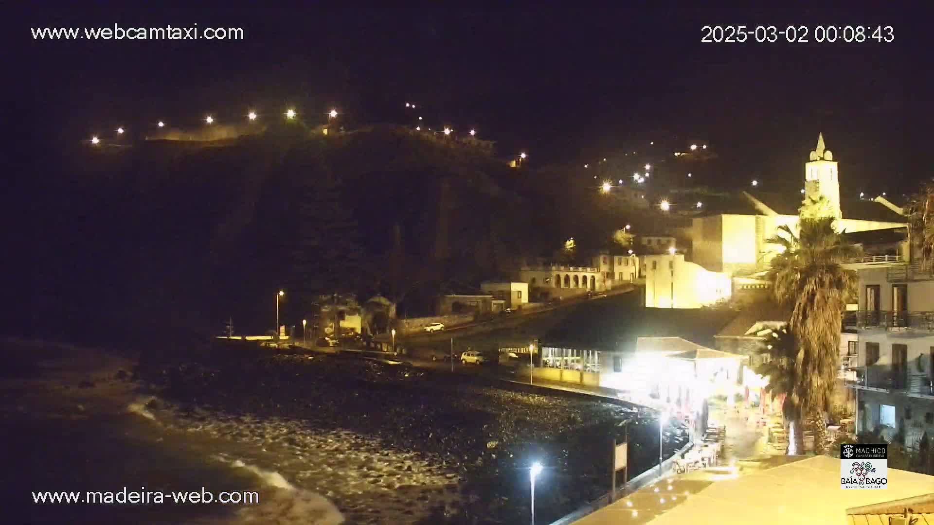 A nighttime coastal town scene shows a rocky shoreline with gentle waves, illuminated buildings on a hillside, and a prominent church tower.