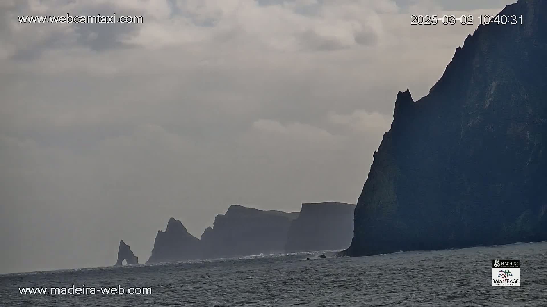 Dark, imposing cliffs rise from a gray, somewhat choppy sea under an overcast sky.