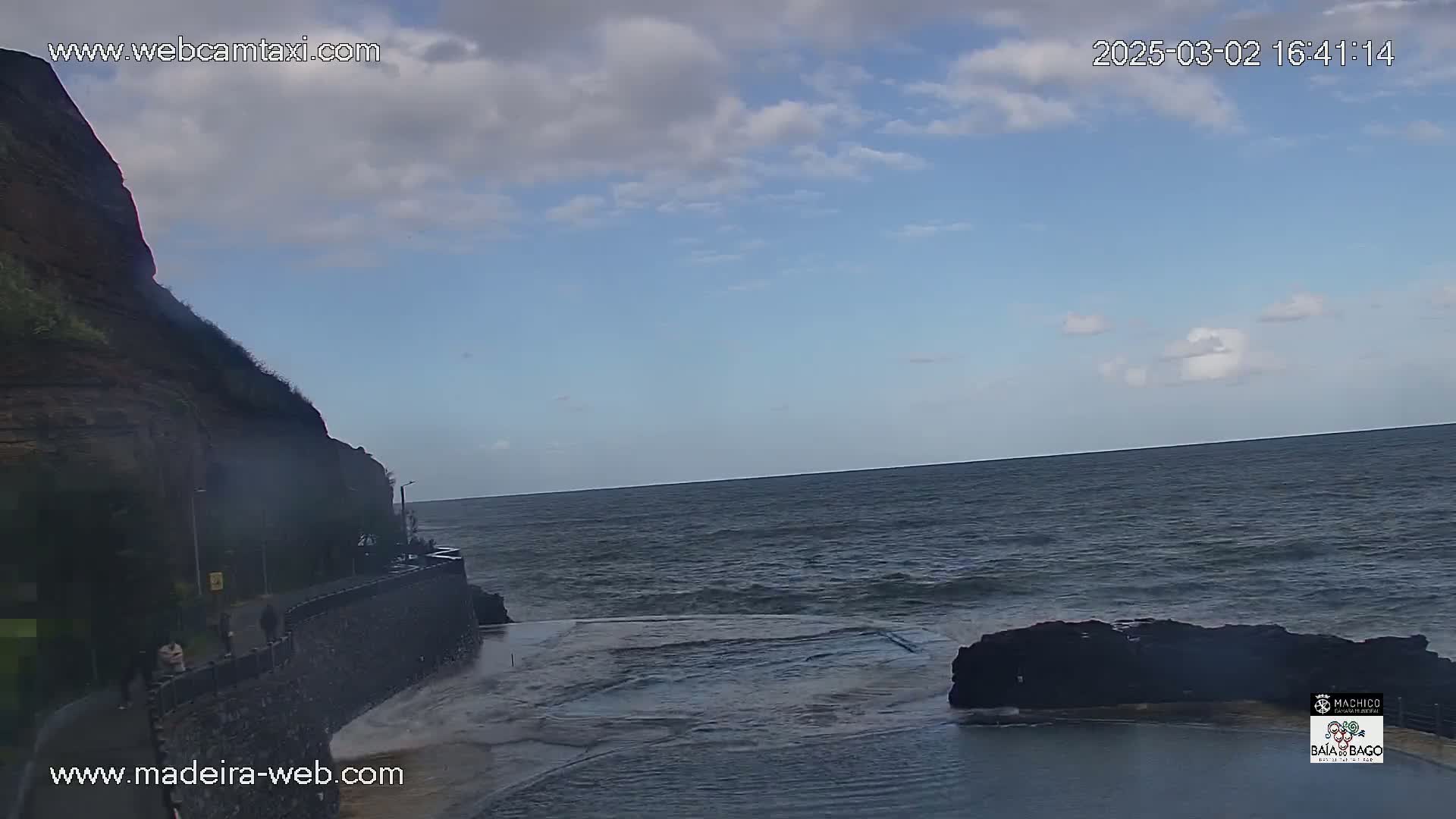 A rocky cliff overlooks a dark-blue ocean with some small waves under a partly cloudy sky.