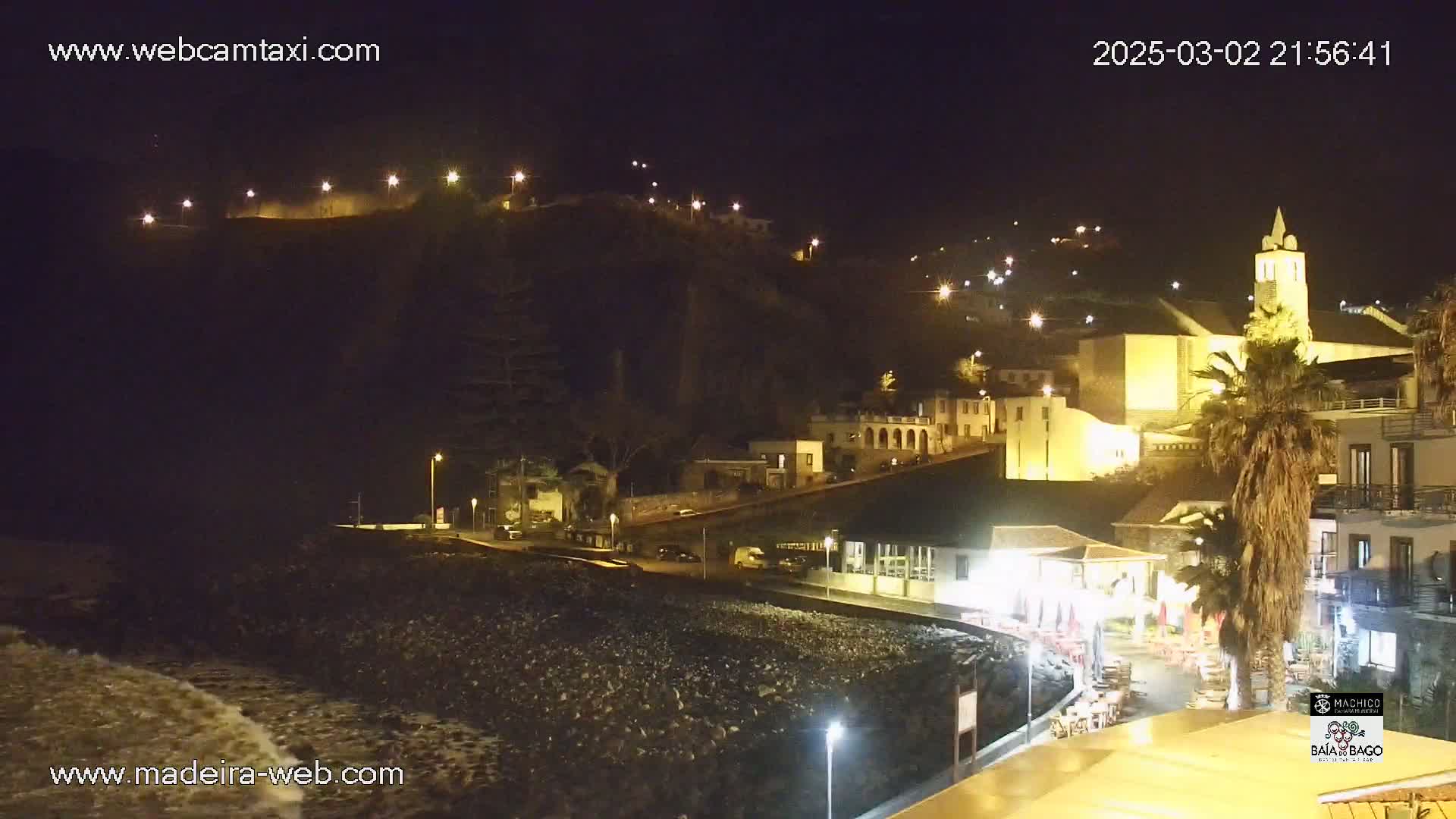 A nighttime view of a coastal town nestled against a hill, illuminated by streetlights, with gentle waves lapping at a rocky shore.