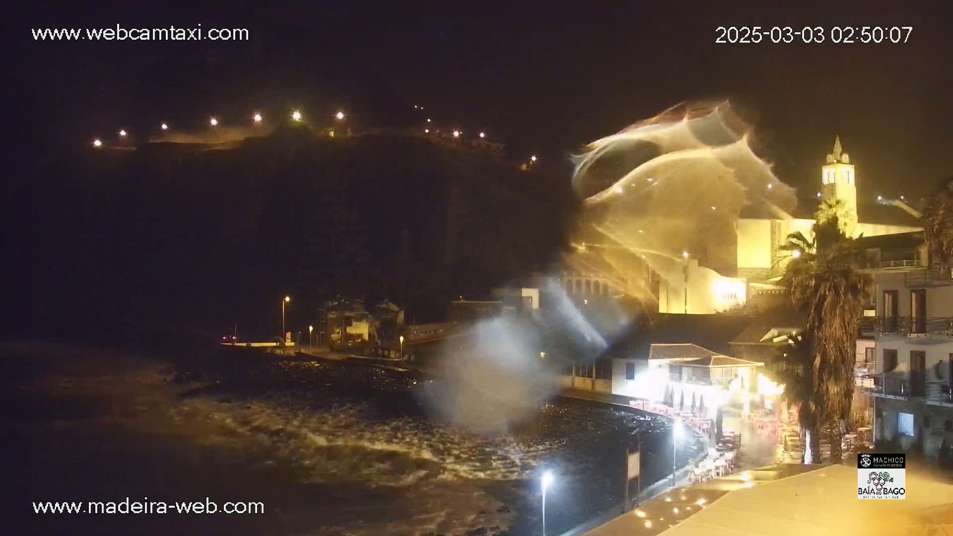 Large waves crash against a shoreline at night, illuminated by streetlights, near buildings and a hill.