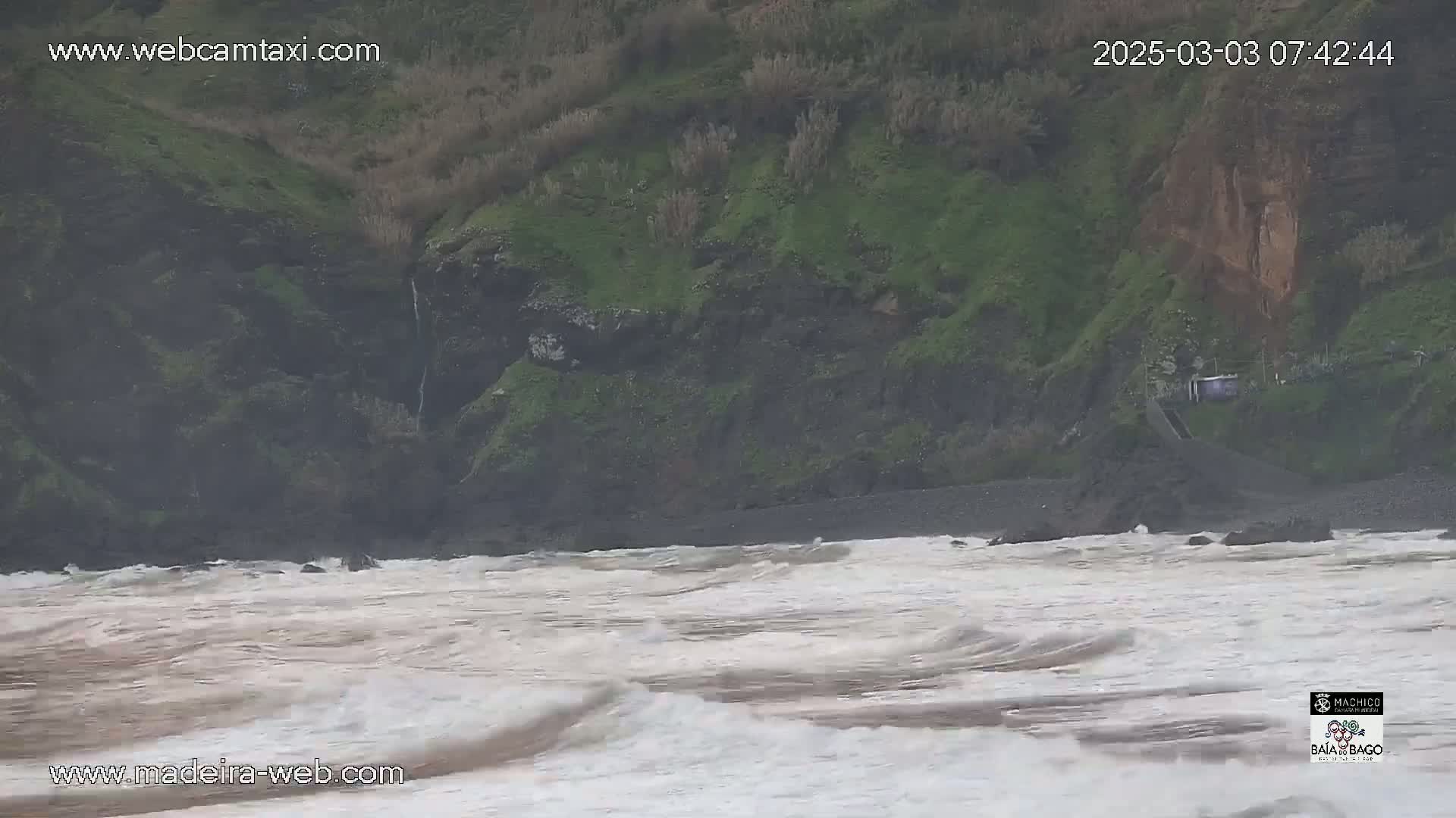 A rocky, green hillside overlooks a choppy, brown-tinged ocean with small waves.