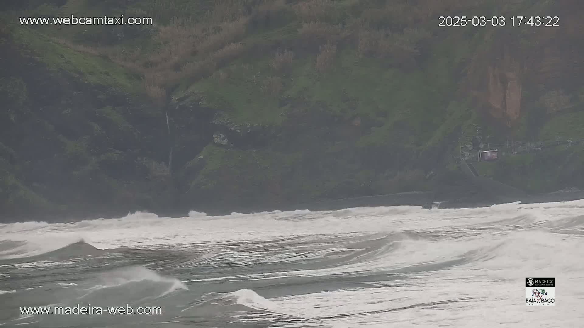 Rough waves crash on a rocky, green shore under an overcast sky.