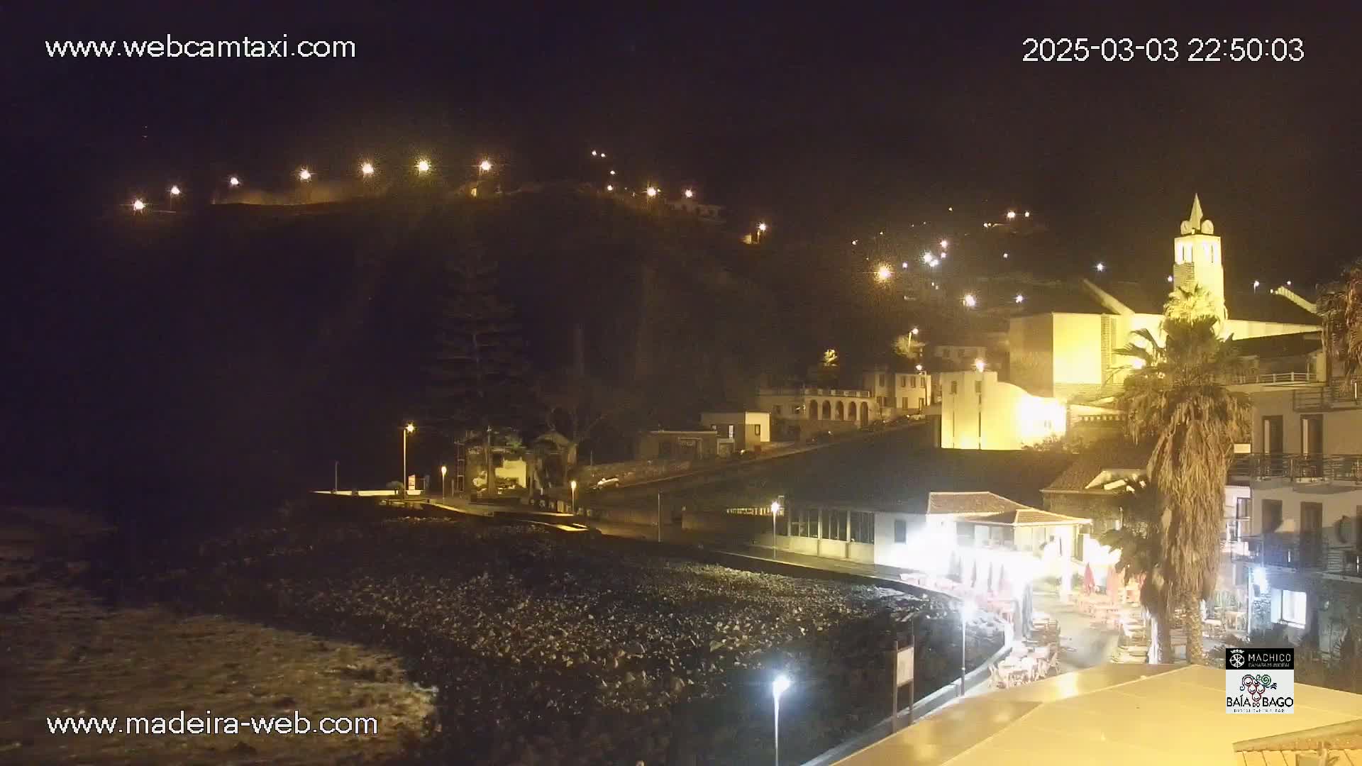 A nighttime coastal town scene shows buildings illuminated against a dark hill under a clear sky.