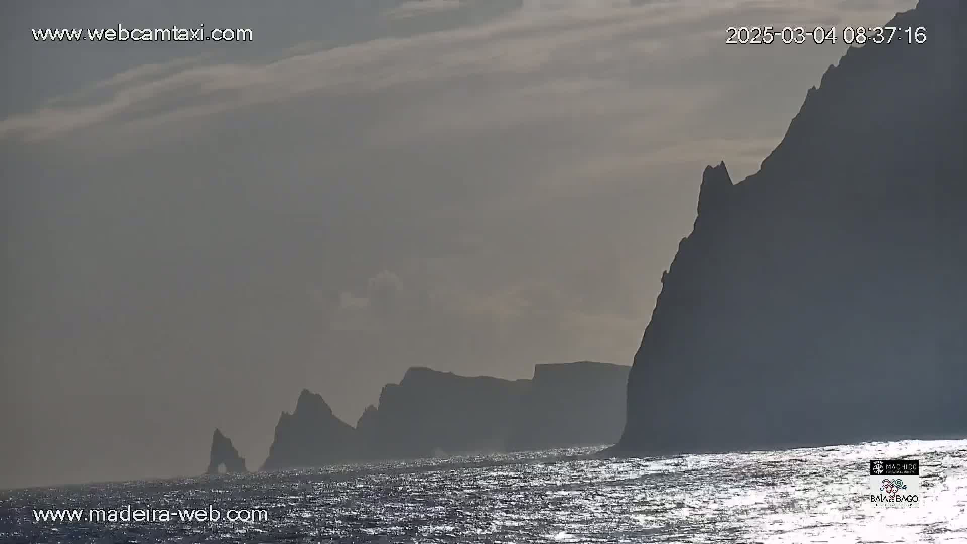 Dark, hazy cliffs rise from a slightly sparkling ocean under a mostly cloudy sky.