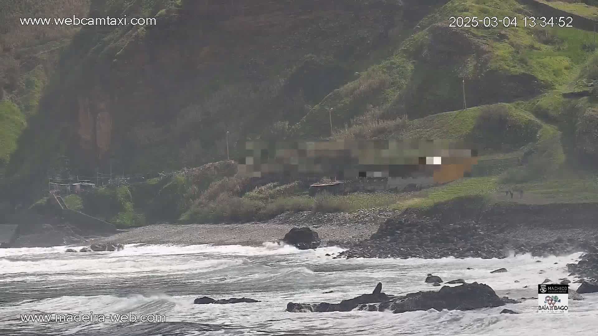 Waves crash on a rocky shore below a green hillside with a few buildings, under a mostly cloudy sky.