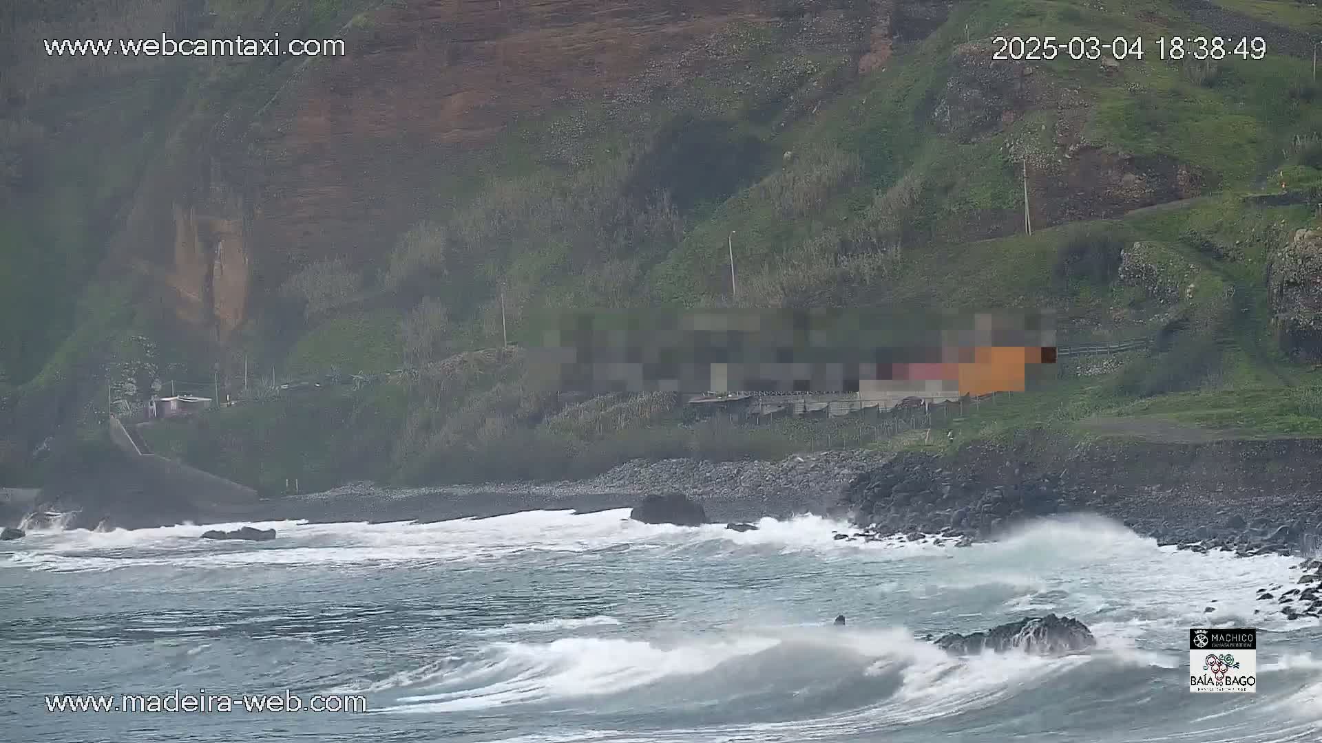 Rough waves crash against a rocky shoreline near a small building situated on a grassy hillside.
