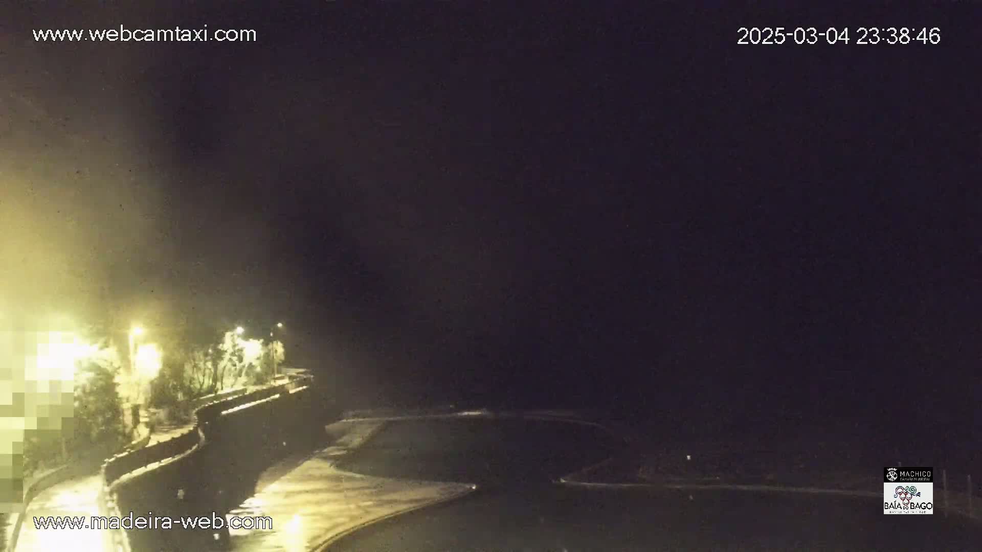 A nighttime coastal scene shows a dark, possibly overcast sky over a paved walkway curving along a shoreline, illuminated by streetlights.