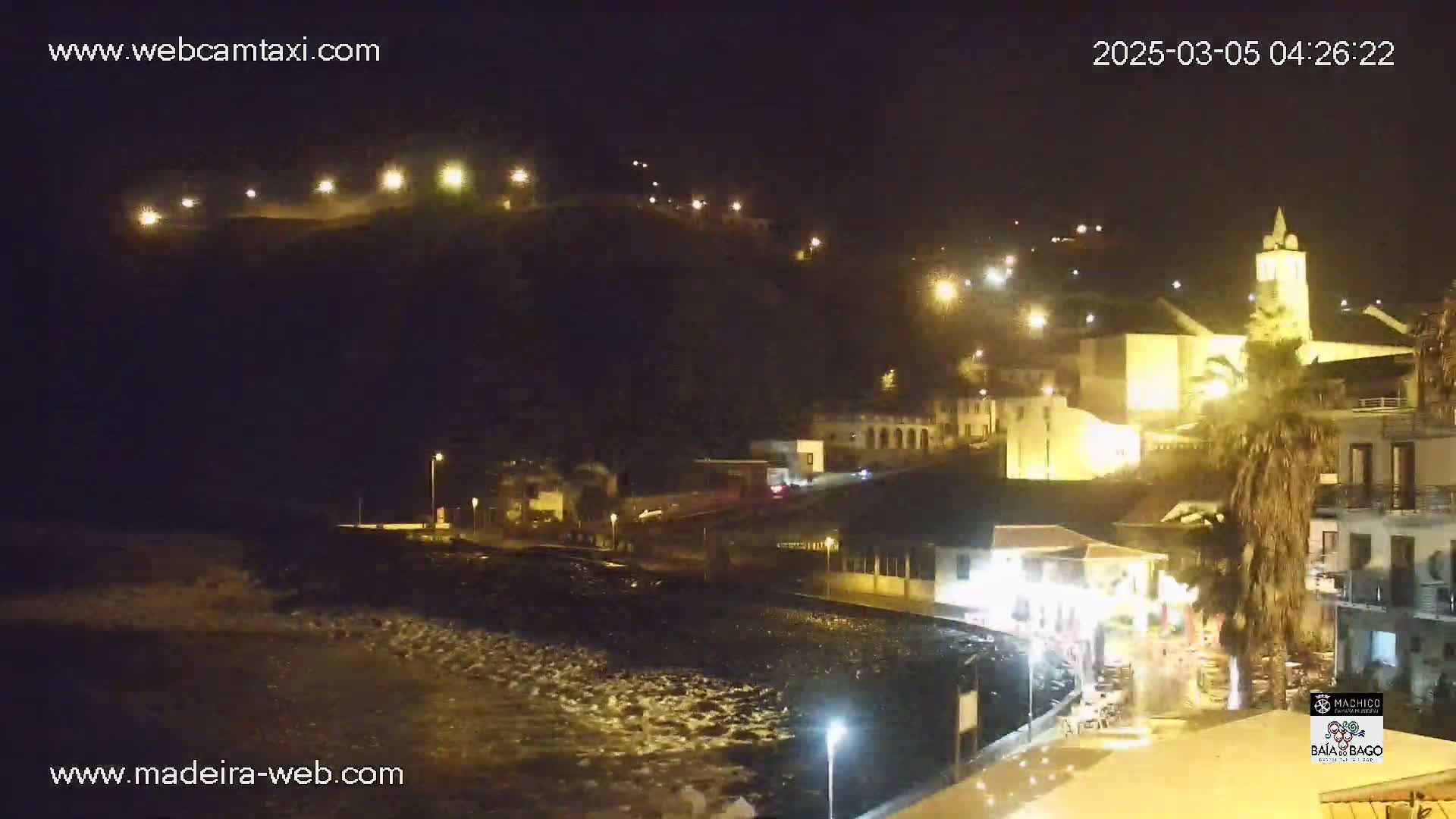 A nighttime view of a coastal town with illuminated buildings and a hilltop, showing a dark, possibly wavy body of water in the foreground.