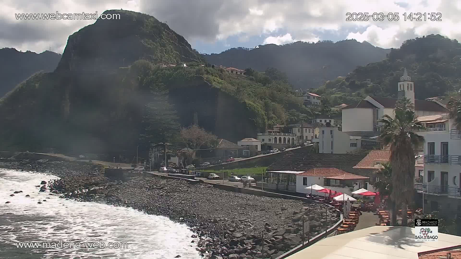 A rocky beach with light waves laps against the shore, next to a small town nestled against a steep, green mountainside under a partly cloudy sky.