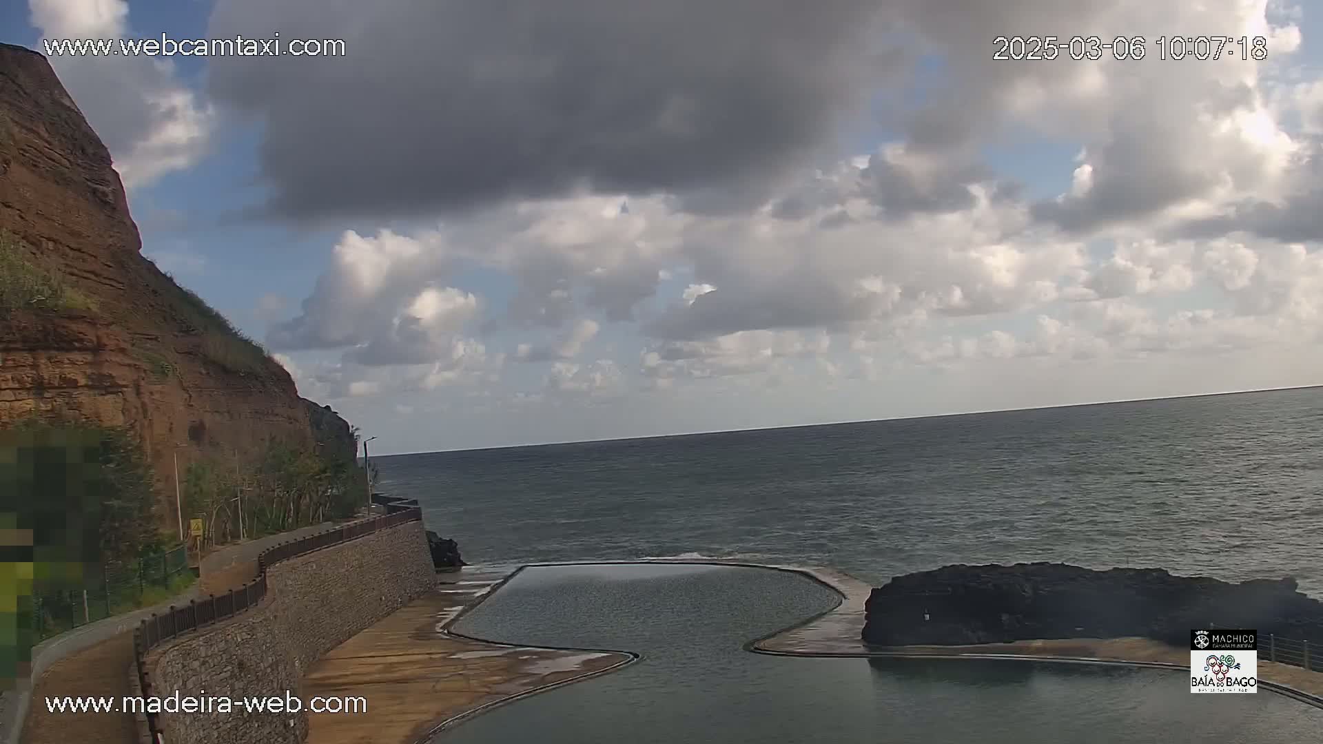 A partly cloudy sky overlooks a rocky coastline with a natural seawater pool; the ocean is calm.